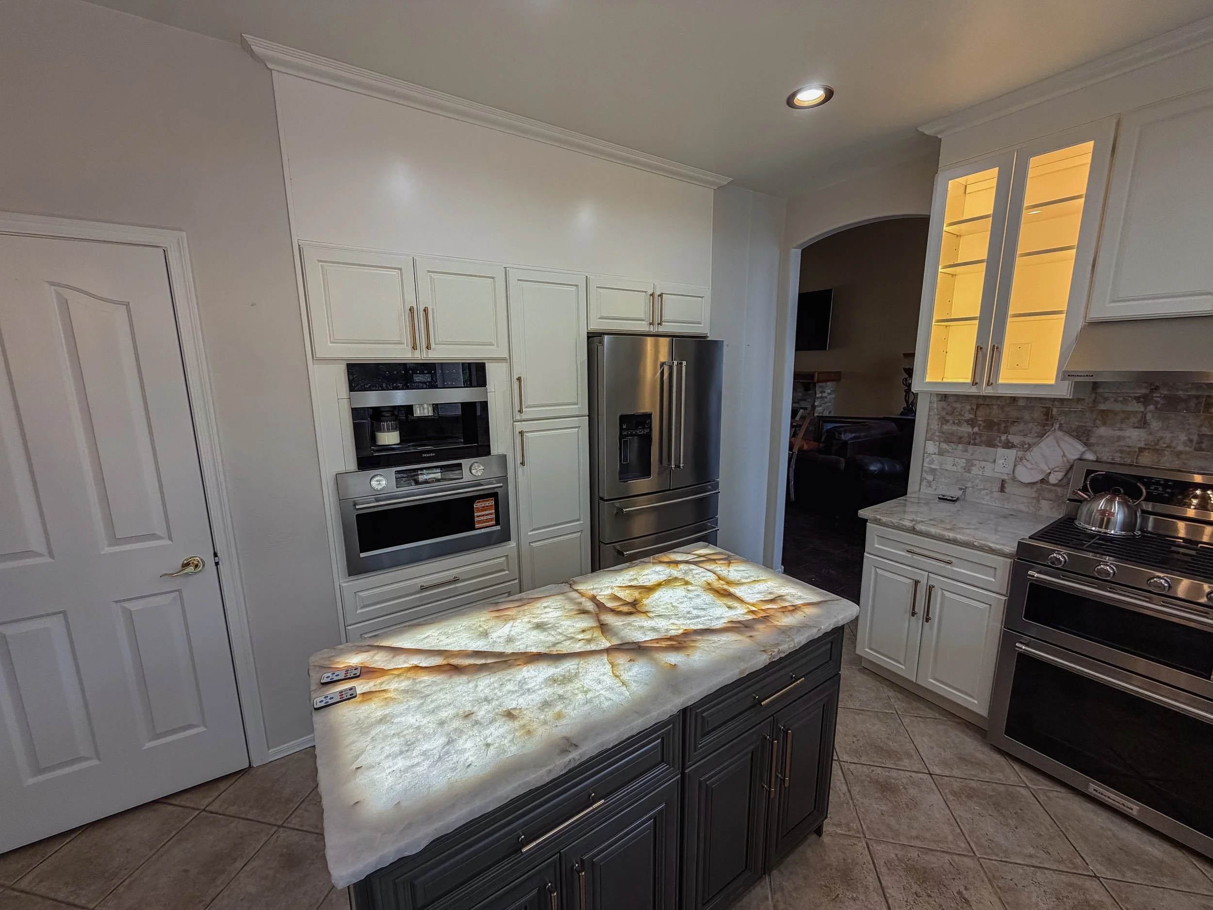 The image shows a modern kitchen with white cabinets, stainless steel appliances, a granite countertop, and a small kitchen island with a colorful onyx countertop. The kitchen has a recessed ceiling light and a view into a living room area with a dark-colored sofa and a TV on the wall.