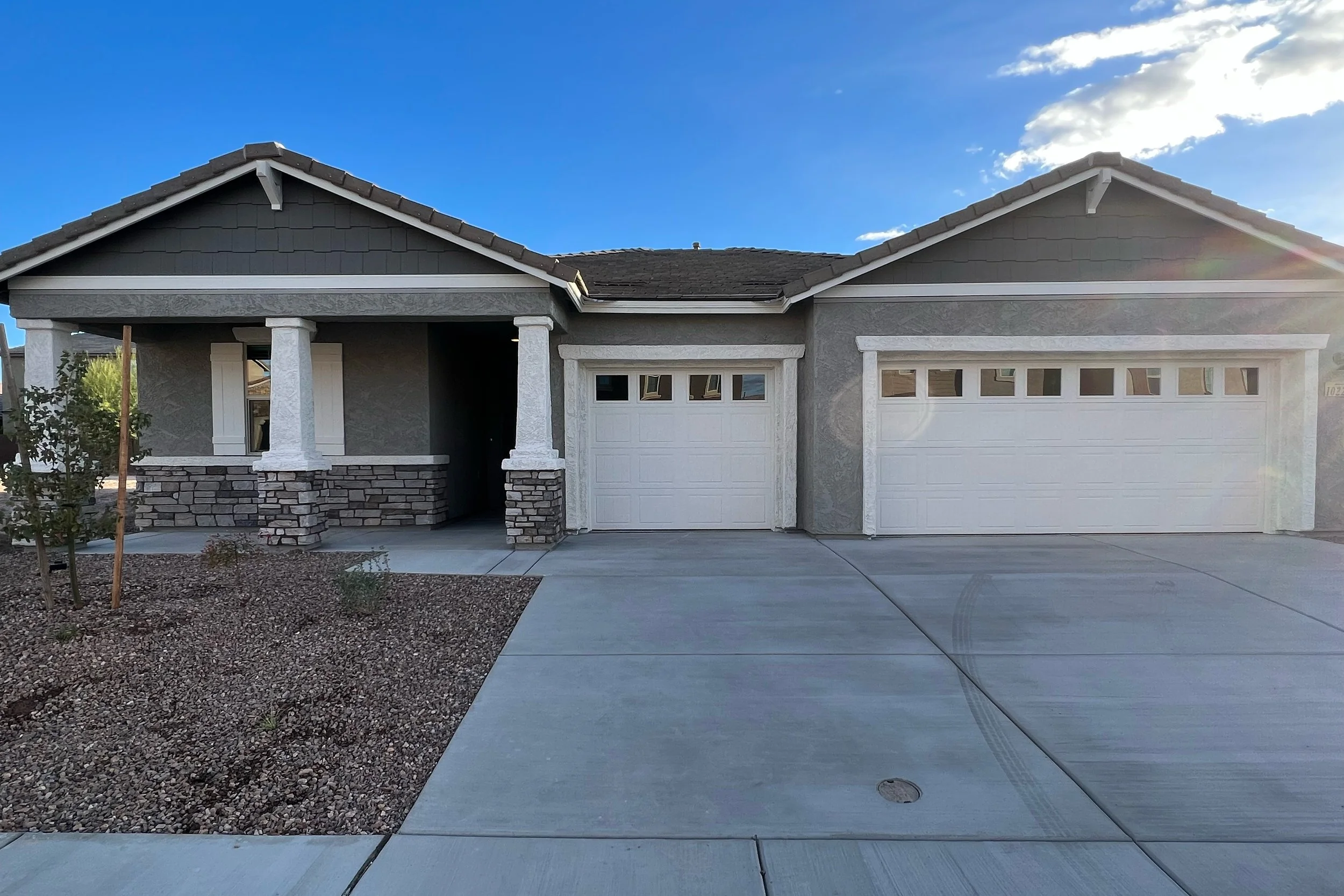 Front view of a modern house with gray exterior walls, two white garage doors, a small covered porch supported by white columns, and a landscaped front yard with small trees and rocks, under a blue sky with some clouds.