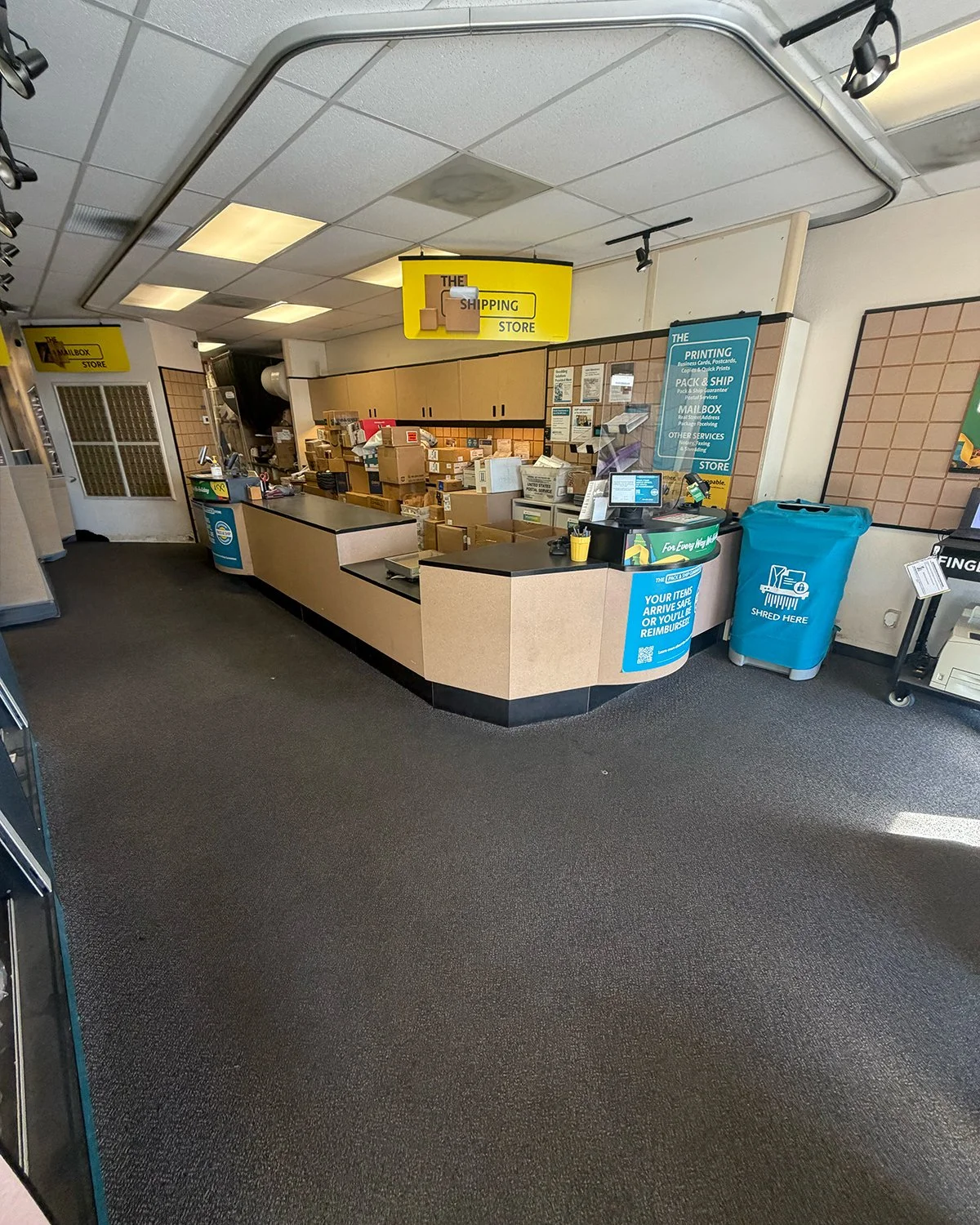 Interior of a shipping store with a counter, stacked packages, and signage for printing, packing, and mailing services.