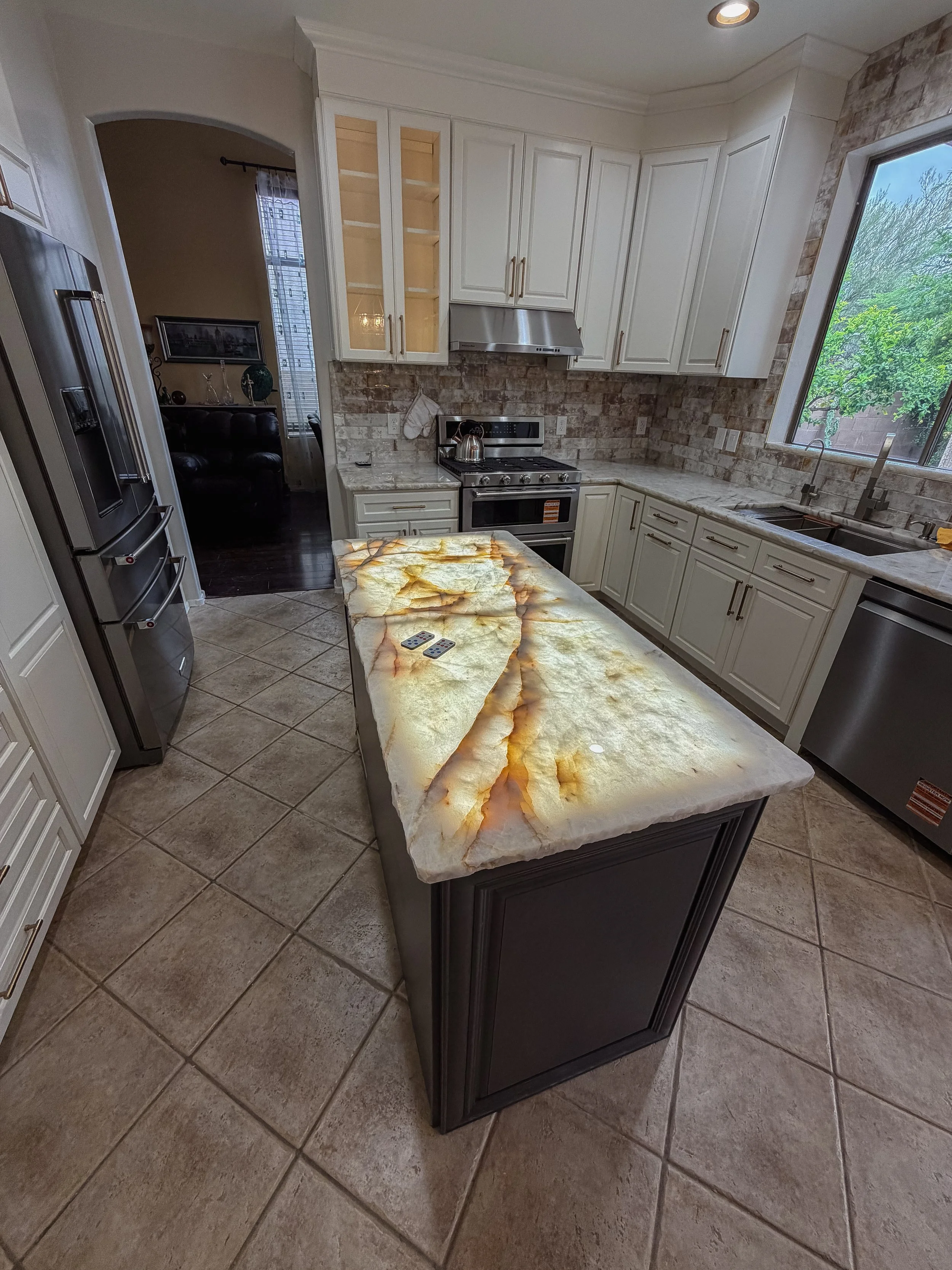 Kitchen with white cabinets, granite countertop, stainless steel appliances, a large window, and tiled floor; counter has remote controls.