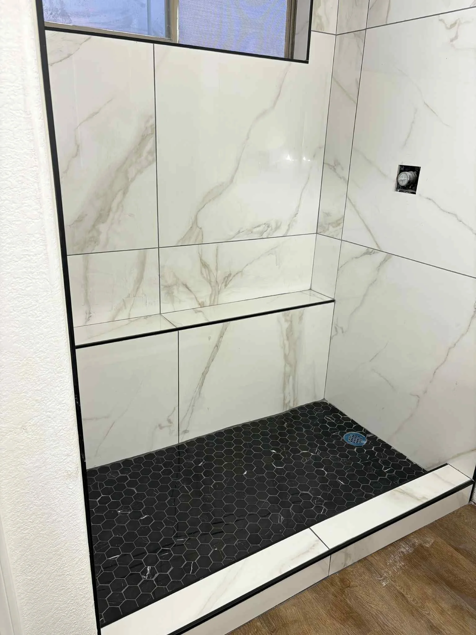 A tiled shower area with large white marble-like tiles on the walls, small black hexagonal tiles on the shower floor, a drain on the floor, and a window at the top.