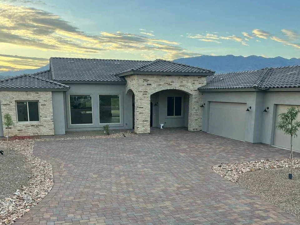 Front view of a new, modern house with an arched entrance, stone and gray siding, and a paved driveway under a sunset sky.