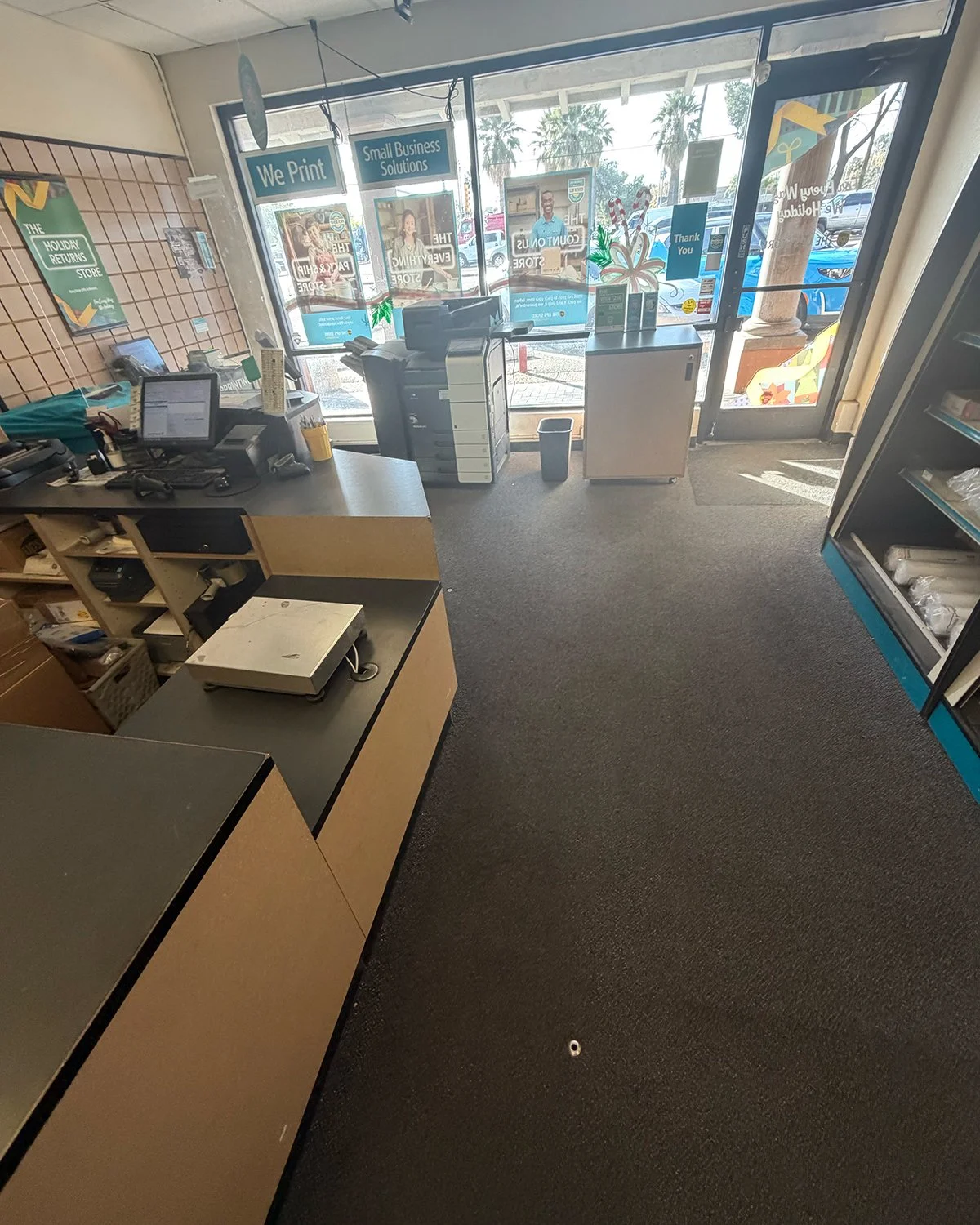 Interior of a store with a checkout counter, computer, printer, and shelves, view through large glass doors with Christmas decorations outside.