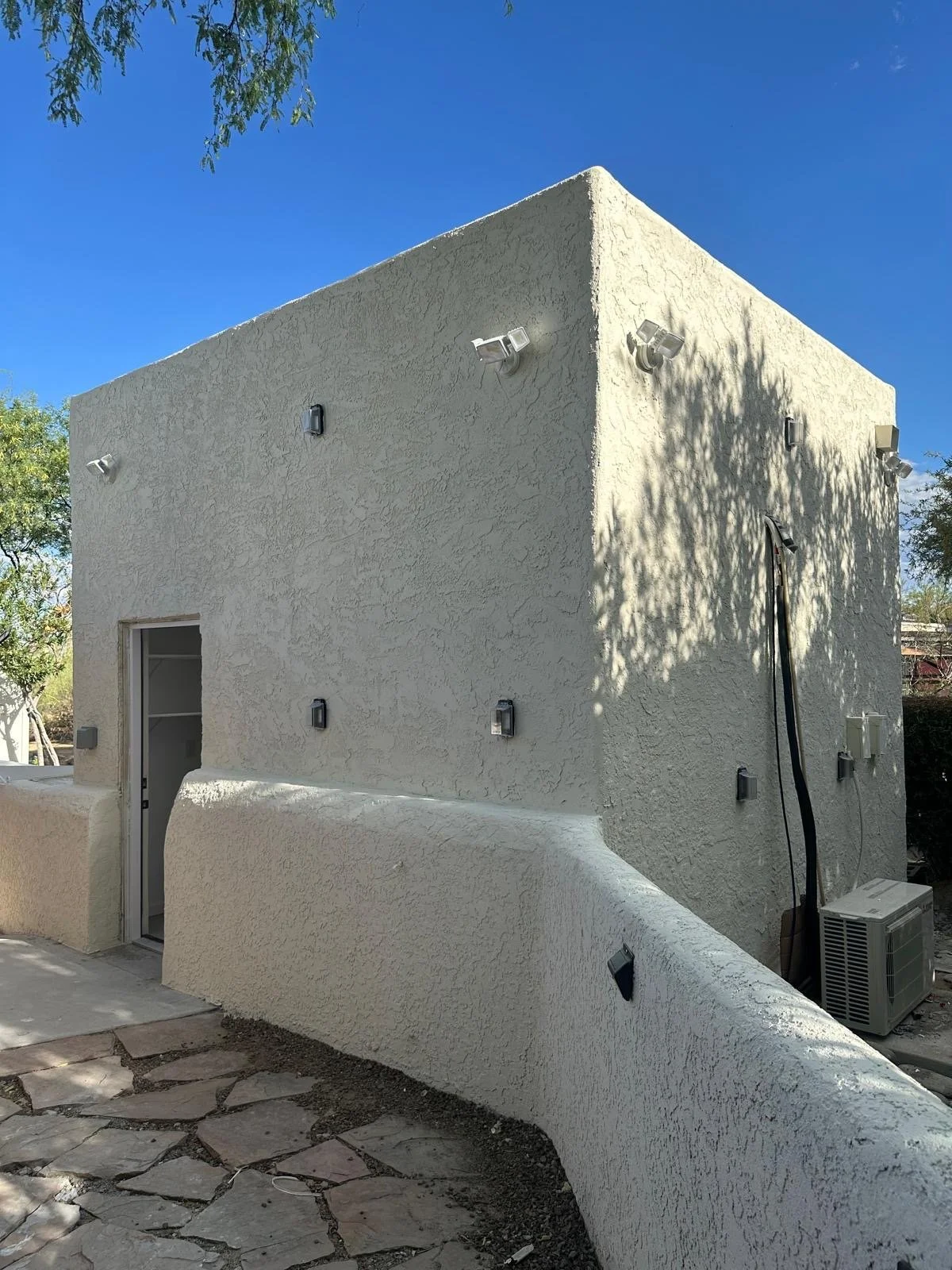 A two-story white stucco building with exterior wall lights and an air conditioning unit outside. Shadows of trees are cast on the wall. The sky is blue with some small clouds.