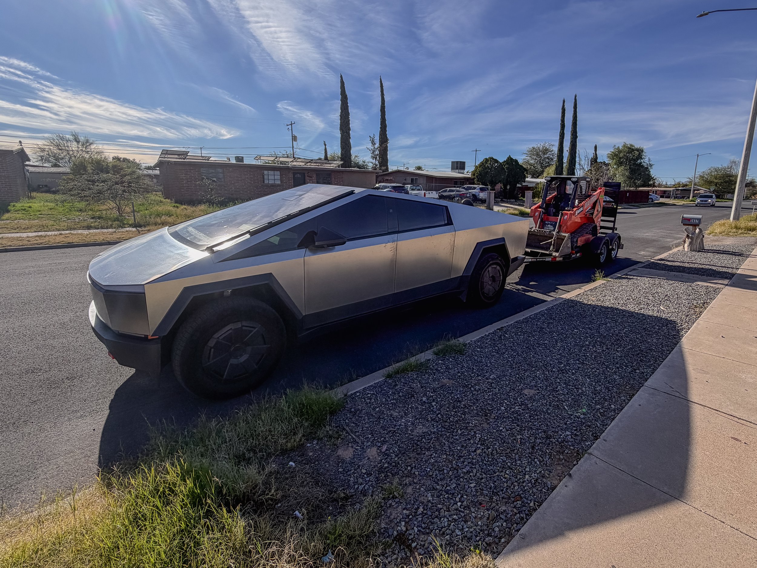 A silver Tesla Cybertruck parked on the street with a small trailer attached. The trailer appears to be carrying equipment or materials. The scene is under a bright blue sky with some wispy clouds, and there are residential houses and trees in the ba