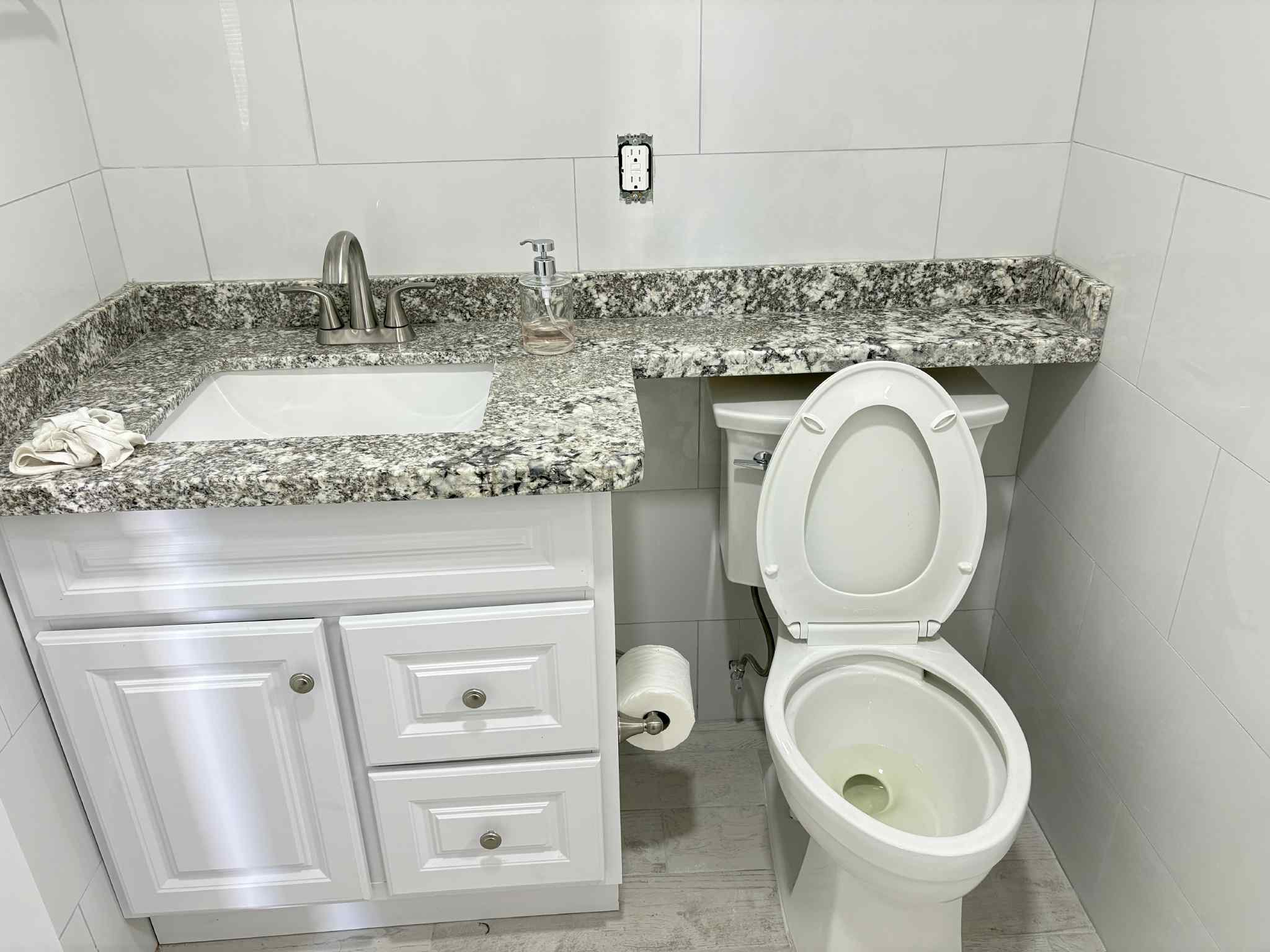 Bathroom with granite countertop, white cabinetry, sink, soap dispenser, toilet with lid open, and wall outlet.