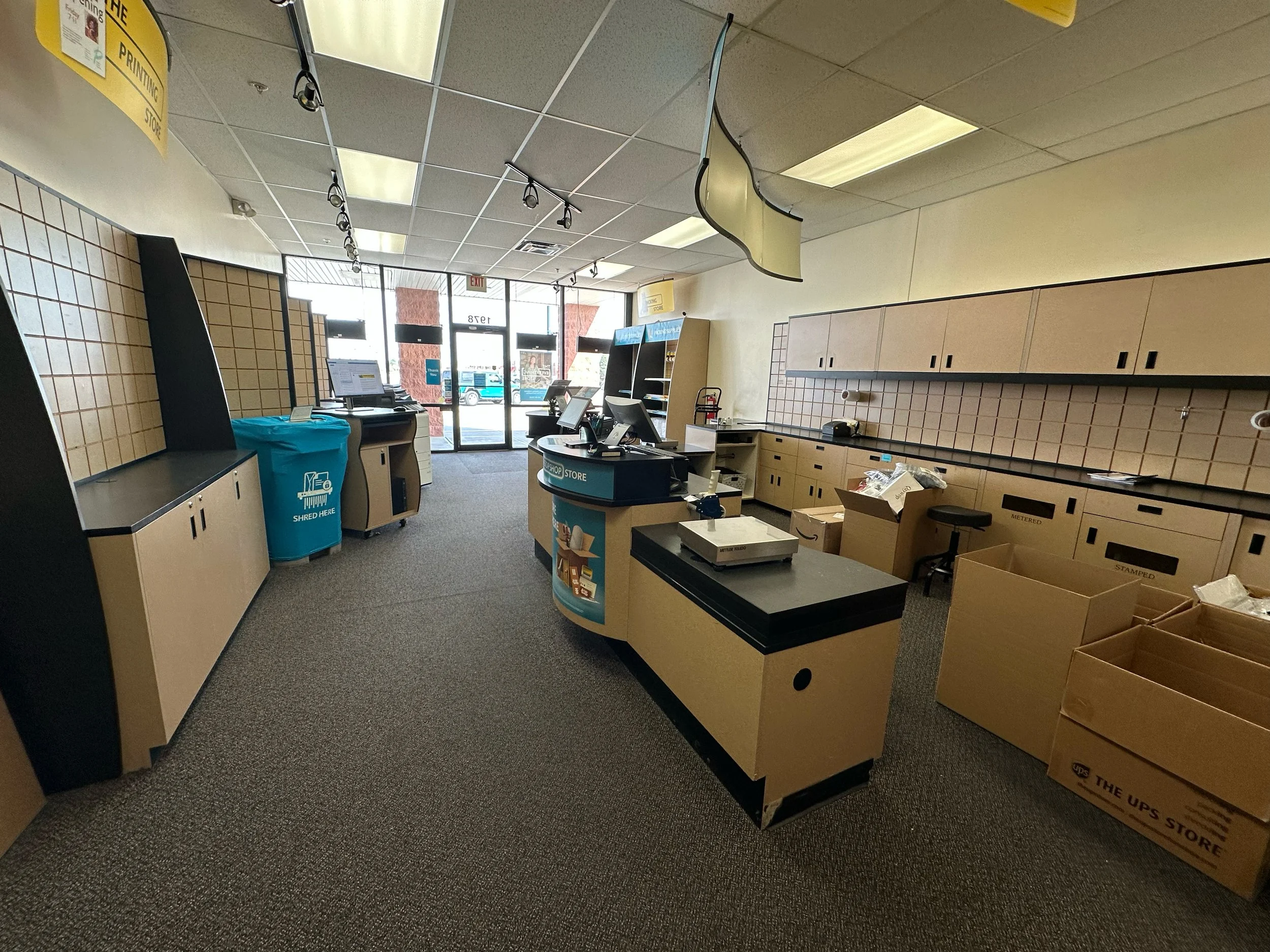 Empty store interior with checkout counters, cardboard boxes, shelving units, and a blue recycling bin near the entrance.