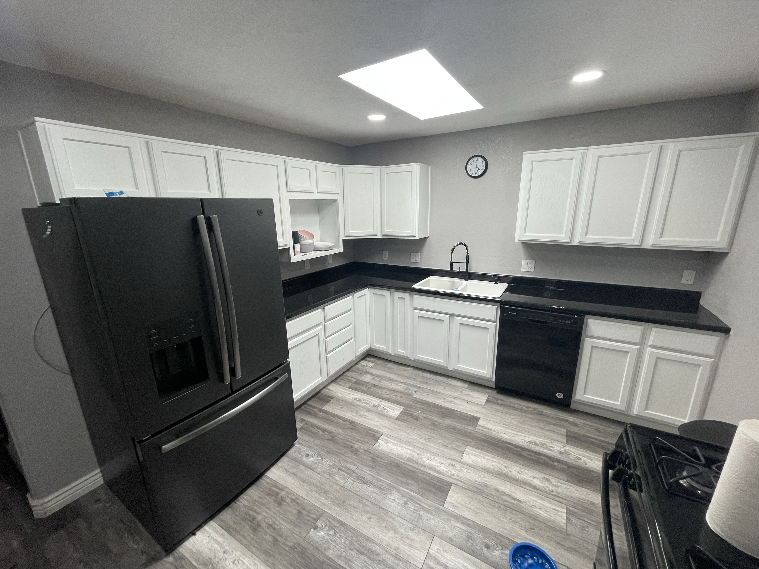 A modern kitchen with white cabinets, black countertops, a stainless steel refrigerator, and wood-look flooring. There is a wall clock and a sink under the cabinets, and a gas stove on one side.