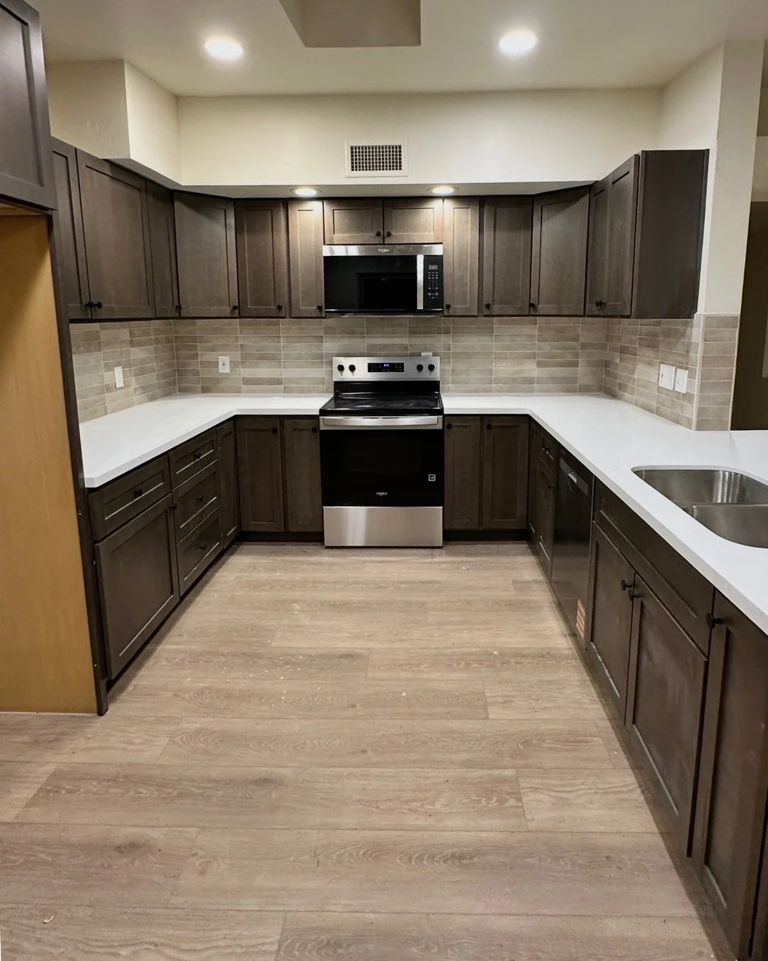 Kitchen with brown cabinets, white countertops, a stove, microwave, and a double sink, with wood flooring and beige tile backsplash.