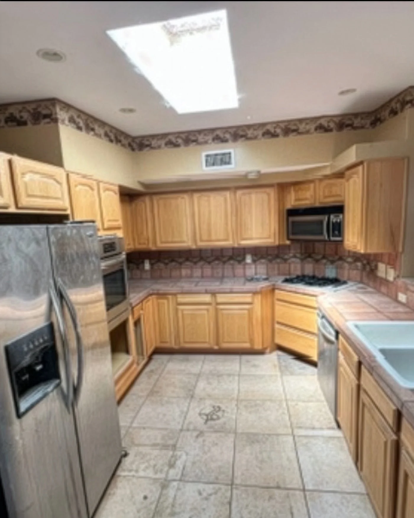 A kitchen with wooden cabinets, stainless steel appliances including a refrigerator, microwave, and oven, tiled countertops, a double sink, and tiled floor with decorative design.