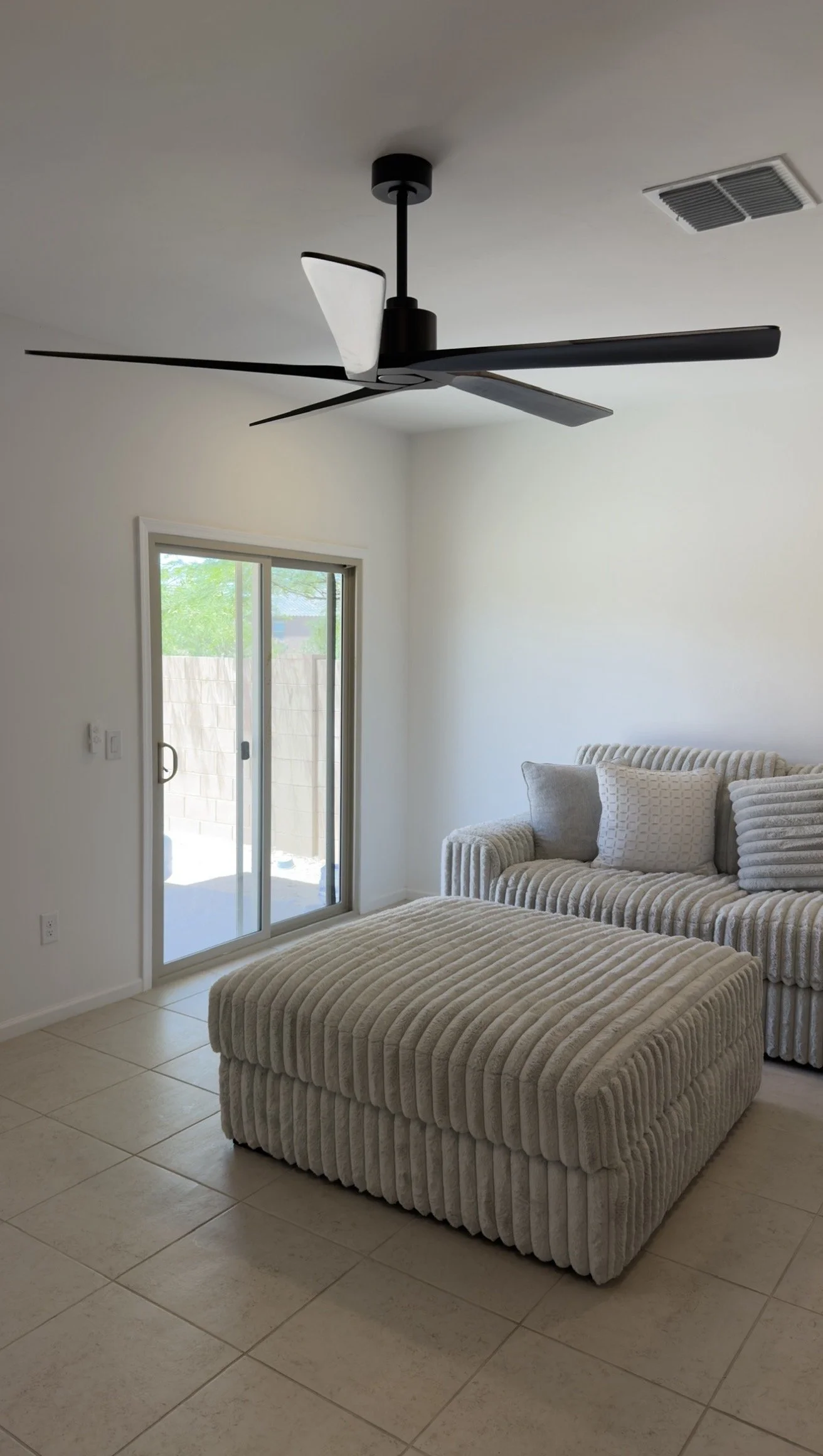 Living room with beige tiled floor, white walls, a sliding glass door leading outside, a plush beige armchair with a matching ottoman, and a contemporary black ceiling fan.