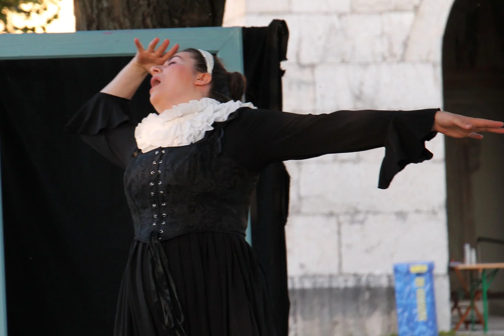 Eine Schauspielerin in historischem Kostüm agiert auf einer Freilichtbühne. Sie trägt ein langes schwarzes Kleid mit Korsett-Schnürung, weiten Ärmeln und eine auffällige, weiße Halskrause. Ihre Haare sind zu einem Dutt hochgesteckt und mit einem weiß