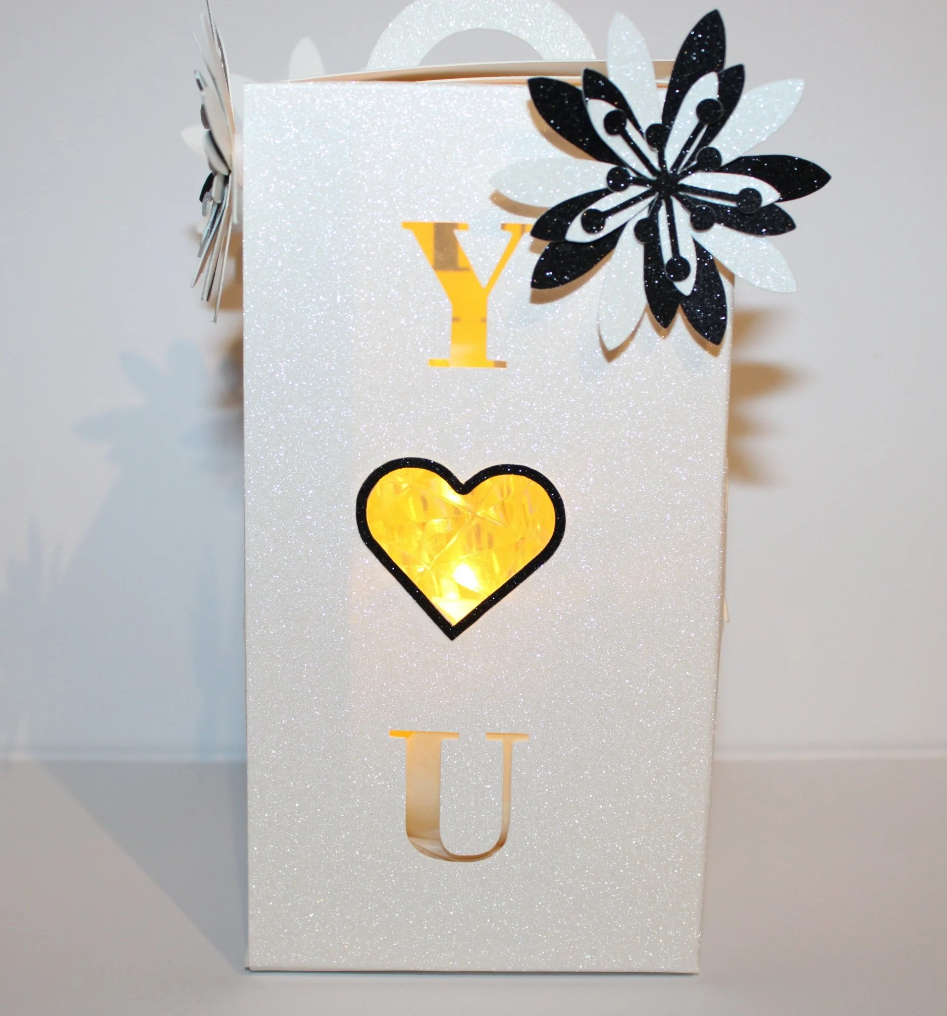 Delicate paper flowers adorn each corner, hand-assembled for a charming, three-dimensional effect. Finished with a soft silk ribbon tied around the closing. A small battery-operated candle is included; acetate windows on the panels let the warm glow 