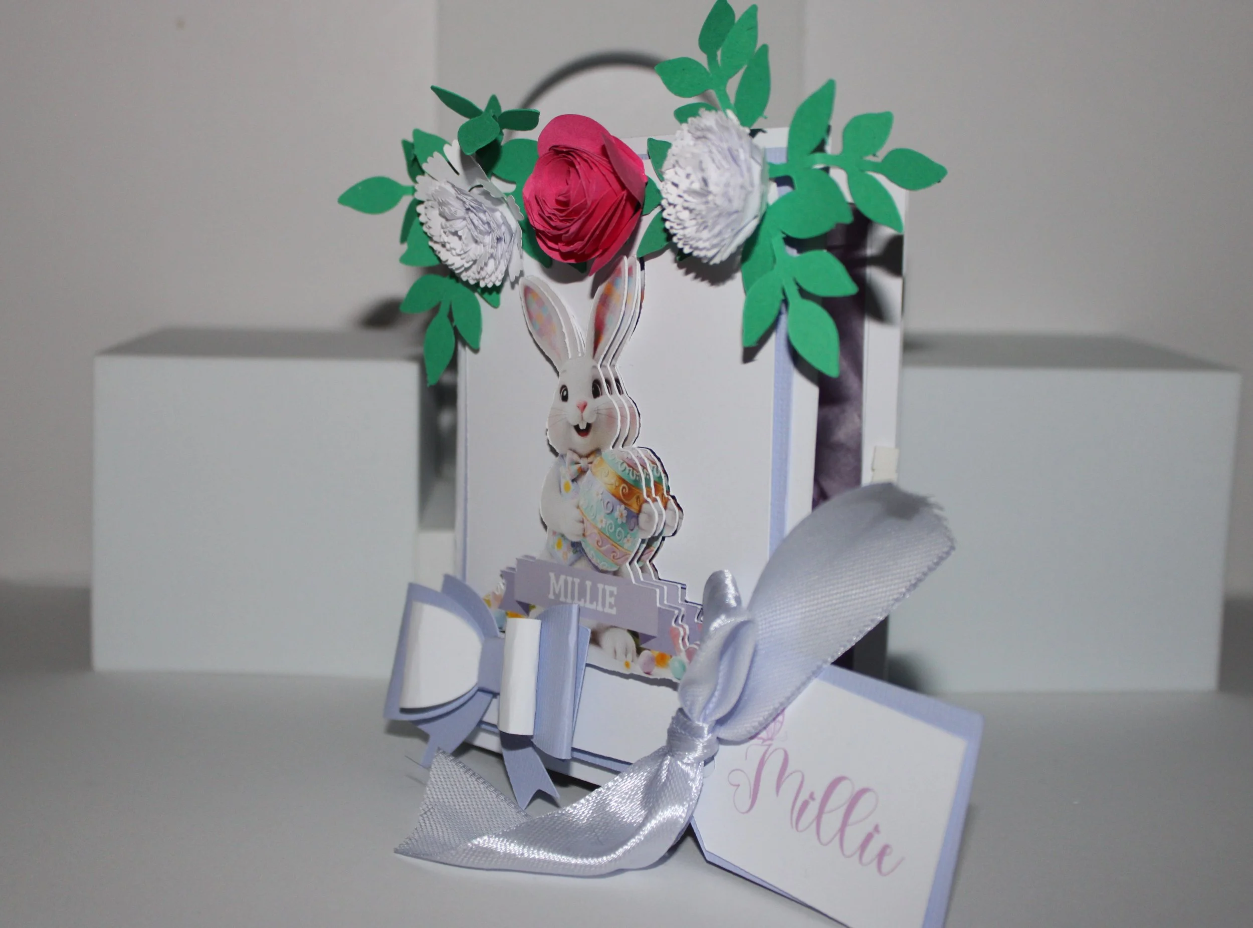Delicate paper flowers cluster in the top corners, while a neat bow and a satin ribbon secure the closure. A personalised gift tag is attached for your message. Open the box to reveal a tissue-paper-lined interior, thoughtfully arranged and filled wi