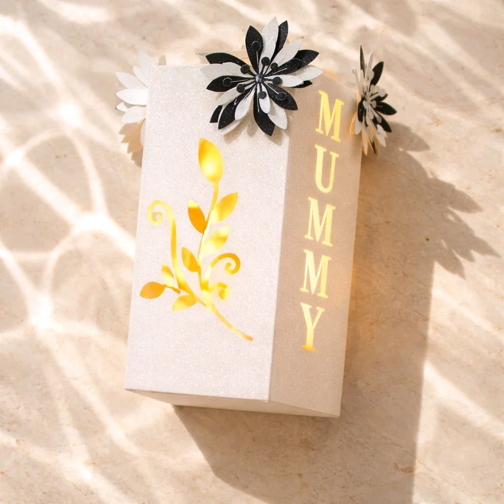 Mothers Day gift box — tall rectangular shape, exquisitely crafted. Each side panel features the words "mummy love you" precision-cut into the box, allowing light to shimmer through. The fourth panel displays a beautifully cut floral motif that compl