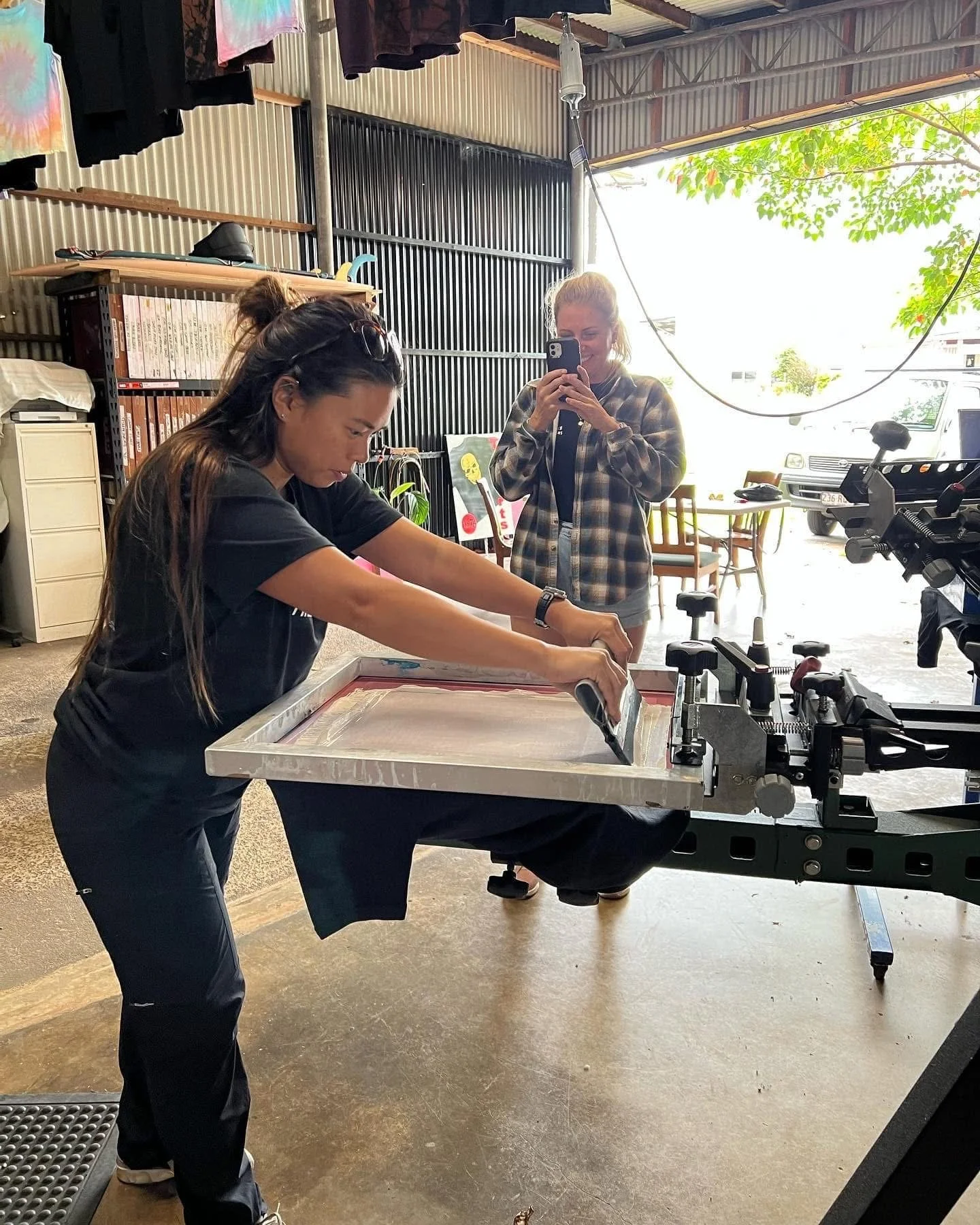 Two women working with a screen printing setup inside a workshop, one is focused on the screen printing process while the other is taking a photo and smiling.