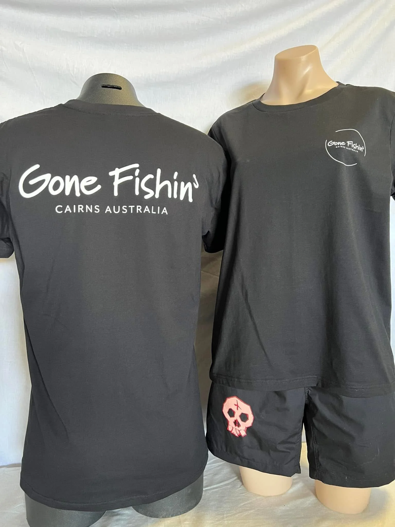 Two black mannequins dressed in black t-shirts and shorts. The mannequin on the left has the text 'Gone Fishin' Cairns Australia' printed on the back, while the mannequin on the right has a small logo with the same text on the front and a skull patch