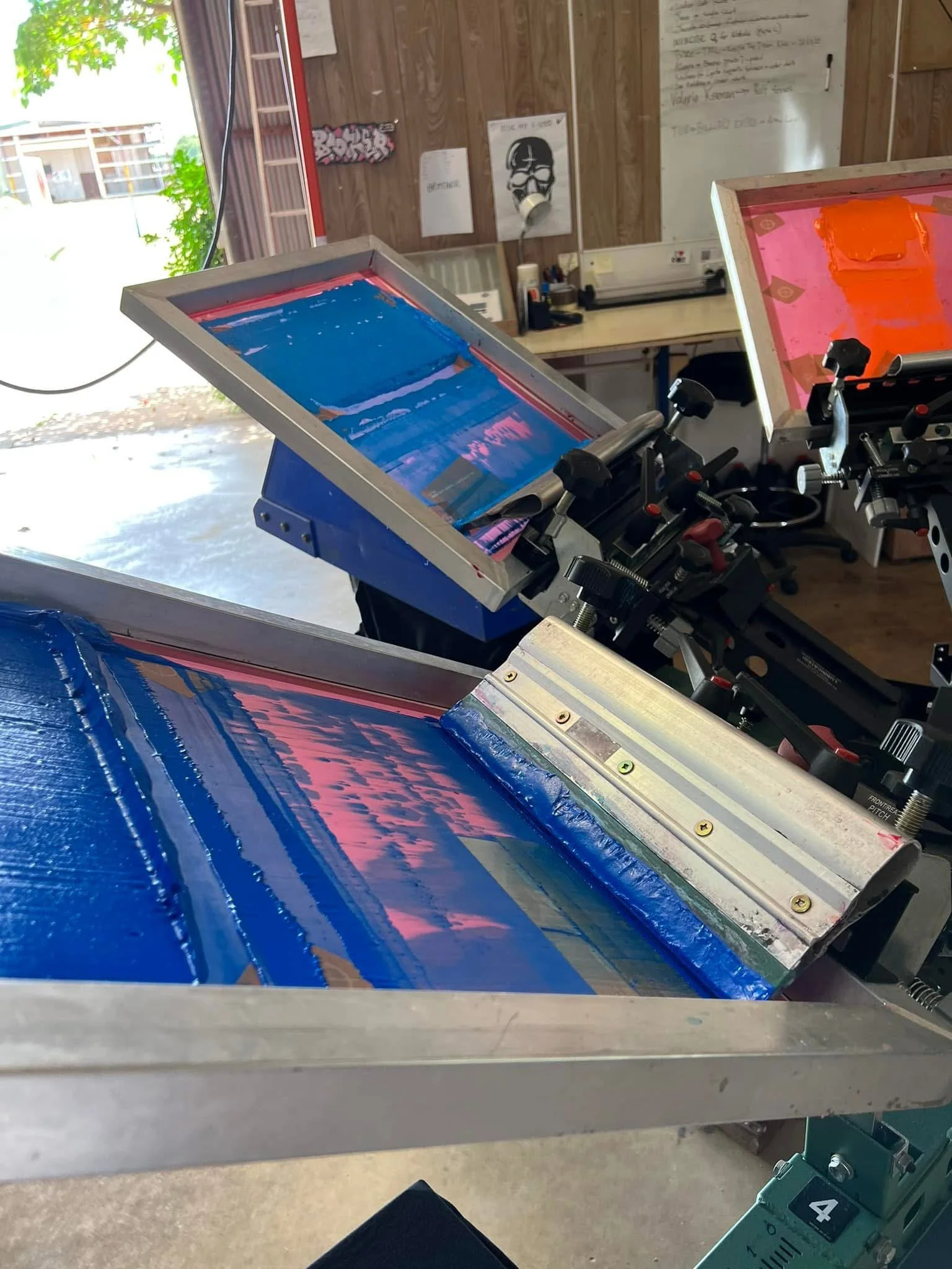 Silkscreen printing presses with pink and blue inks in a workshop. Inks are spread evenly on screens used for printing designs on fabric or paper.