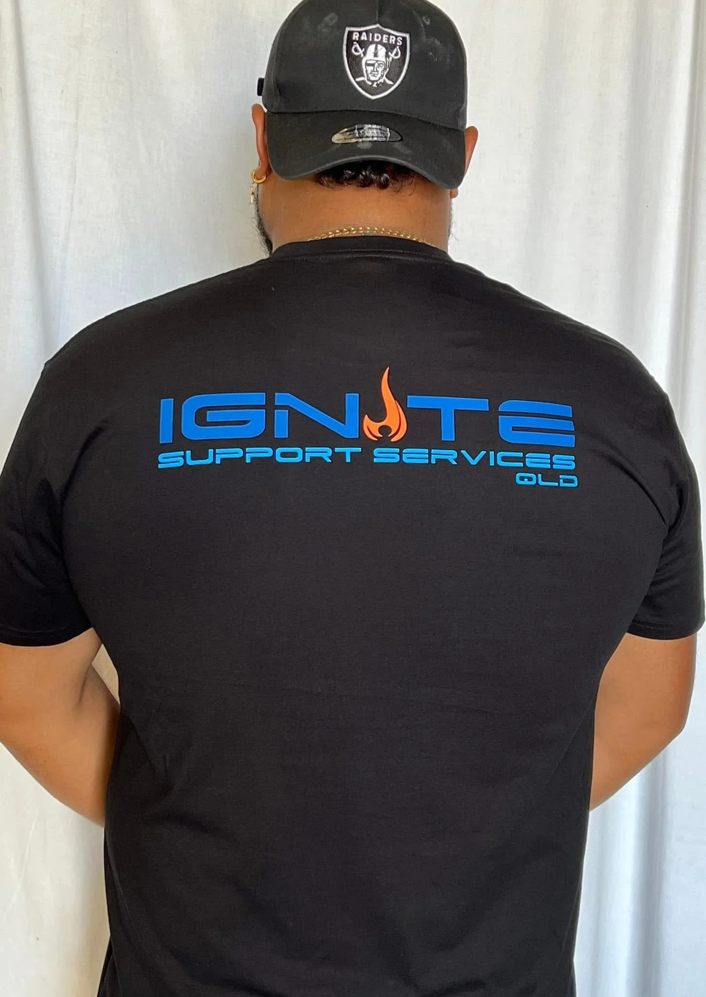 Back of a person wearing a black T-shirt with blue and orange text that reads 'IGNITE SUPPORT SERVICES OLD' and a Raider's hat.