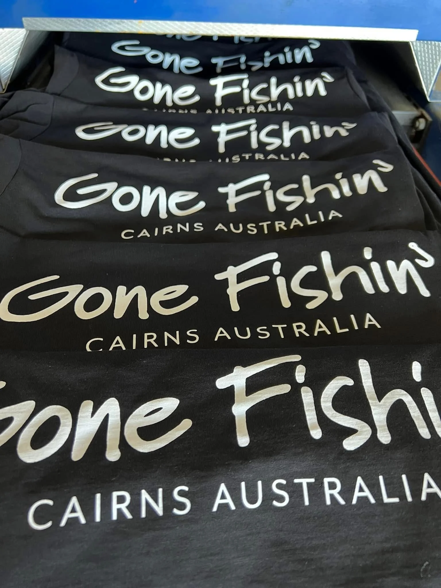 Multiple black T-shirts with white text that reads 'Gone Fishin' CAIRNS AUSTRALIA', folded and stacked in a display.