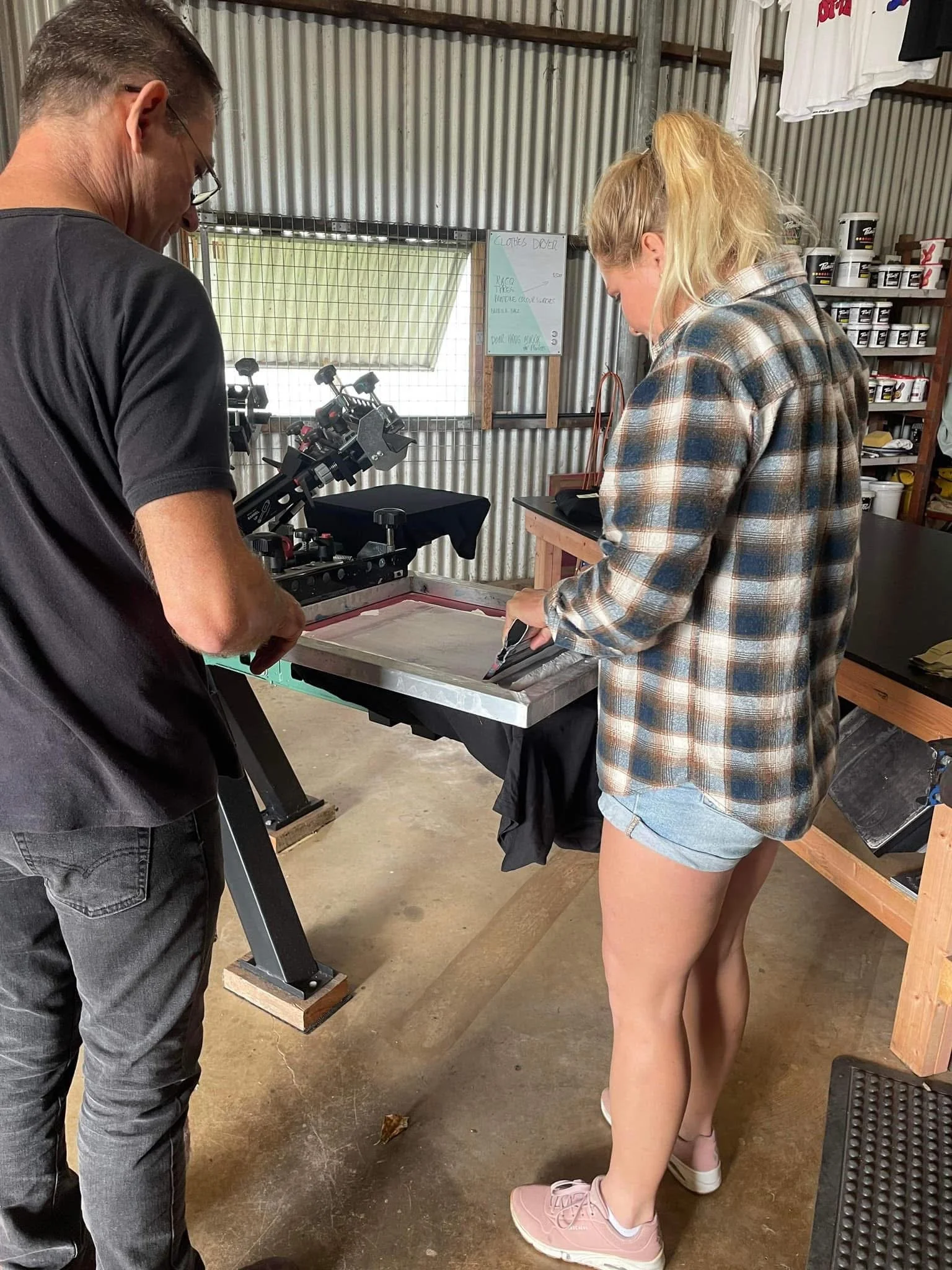 A woman with blonde hair in a ponytail wearing a plaid shirt and shorts uses a screen printing press, while a man in black looks on in a workshop with metal walls and shelves of supplies.