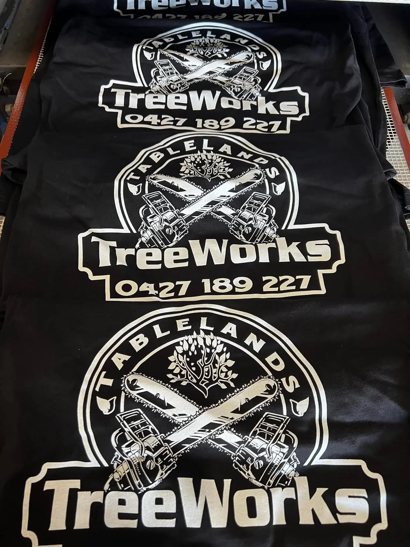 Stack of black T-shirts with a white logo for TreeWorks, featuring crossed chainsaws, a tree, and the text 'TreeWorks' with a phone number.