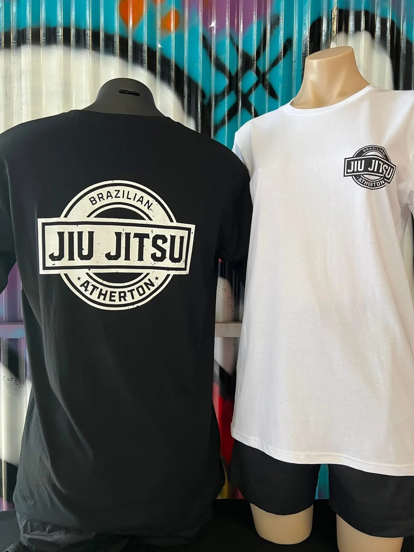 Black and white T-shirts with 'Jiu Jitsu' logo, displayed on mannequins against a graffiti wall.