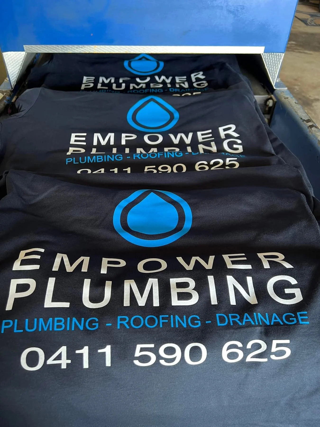 Black T-shirts with blue and white logo and text for Empower Plumbing, displaying services plumbing, roofing, drainage, and phone number 0411 590 625.