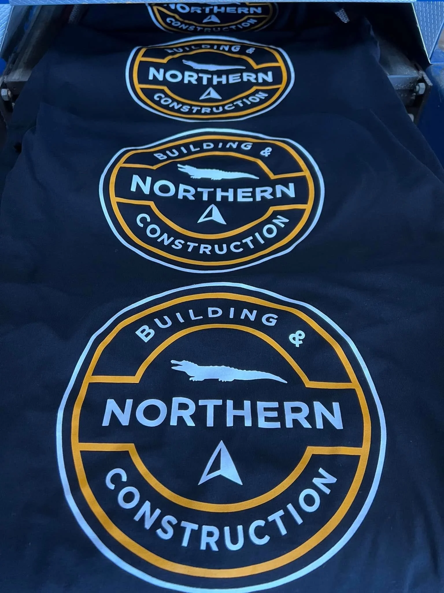 Stack of black items with a circular logo for Northern Building & Construction, featuring a crocodile silhouette, a compass symbol, and yellow, white, and blue text.
