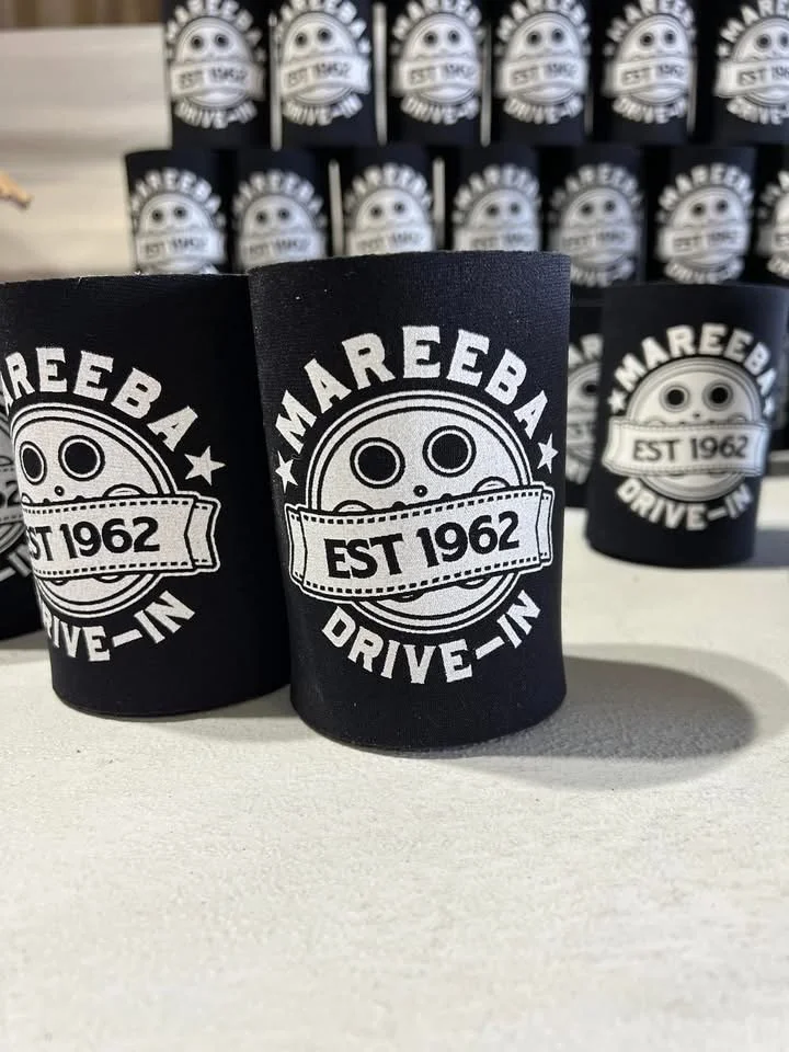 Black insulated mugs with a logo of a smiling face and the text 'Mareeba Drive-In' and 'EST 1962' in white, placed on a white surface with more mugs in the background.