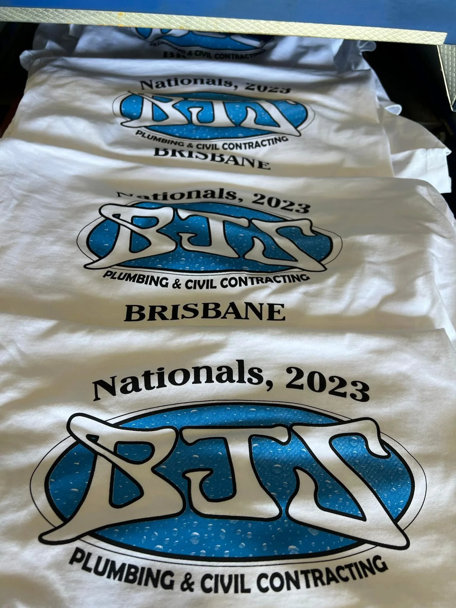 White fabric with a logo for BNT Plumbing & Civil Contracting, Brisbane, for Nationals 2023, laid out flat. Multiple copies of the fabric are visible.