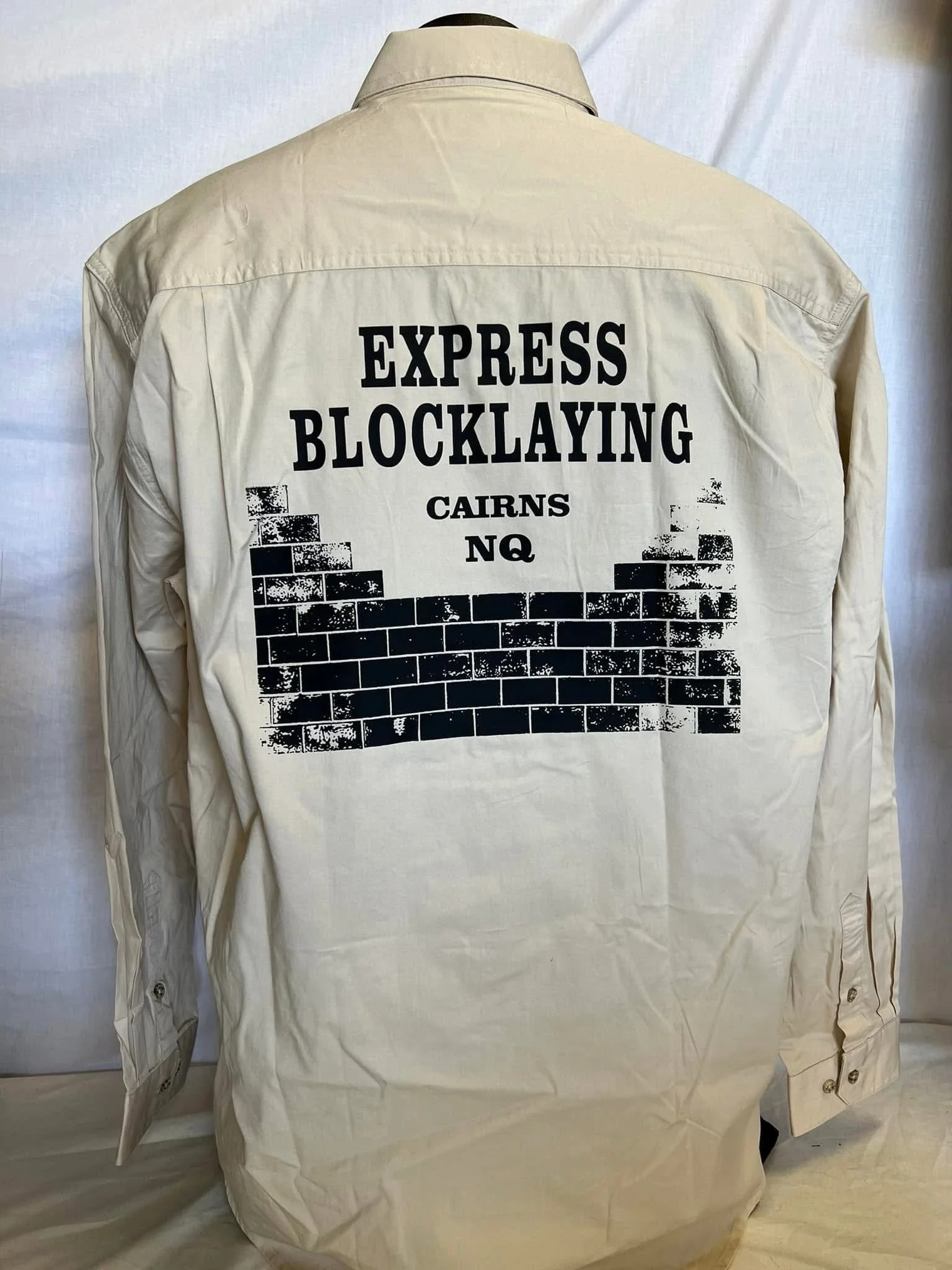 Beige shirt with black printed text on the back that reads 'Express Blocklaying Cairns NQ' and features a graphic of a black brick wall.