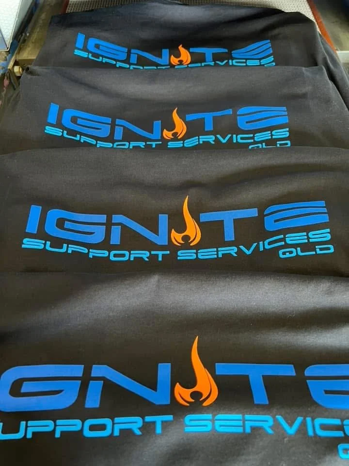 Black T-shirts with blue text reading 'IGNITE SUPPORT SERVICES OLD' and an orange and black flame logo in the center of the word 'IGNITE'.