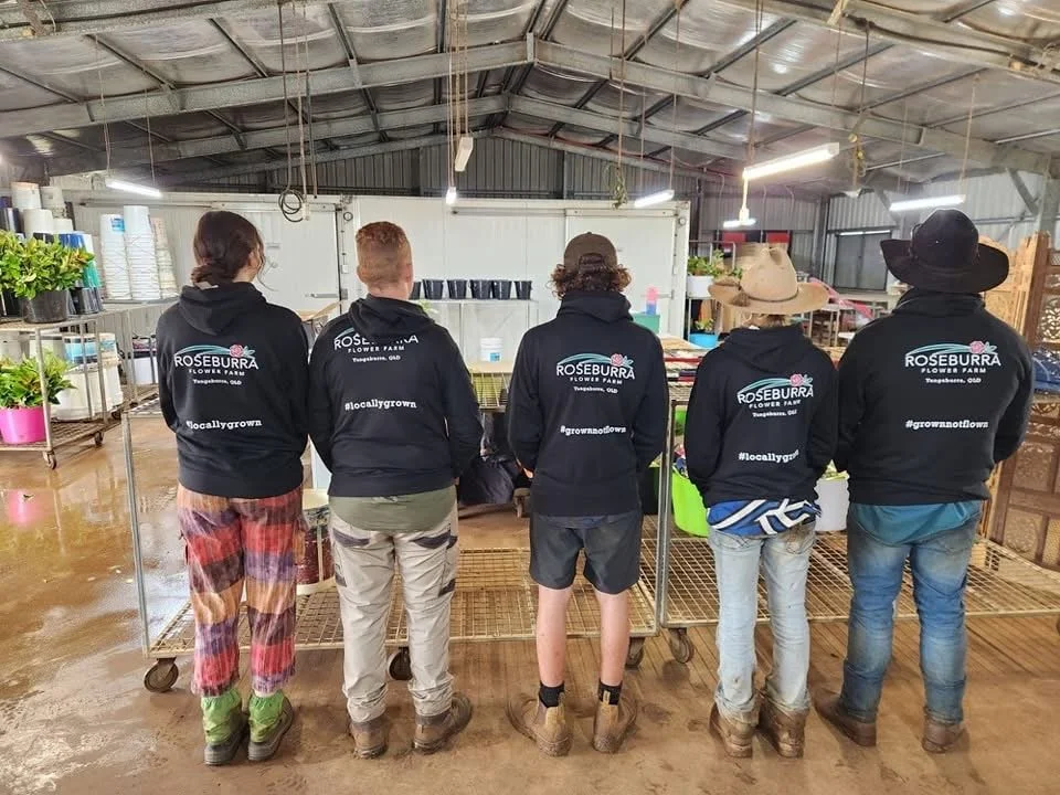 Five people standing in a greenhouse or farm shop, wearing matching black hoodies with 'Roseburra Flower Farm' and #locallygrown printed on the back, facing away from the camera.