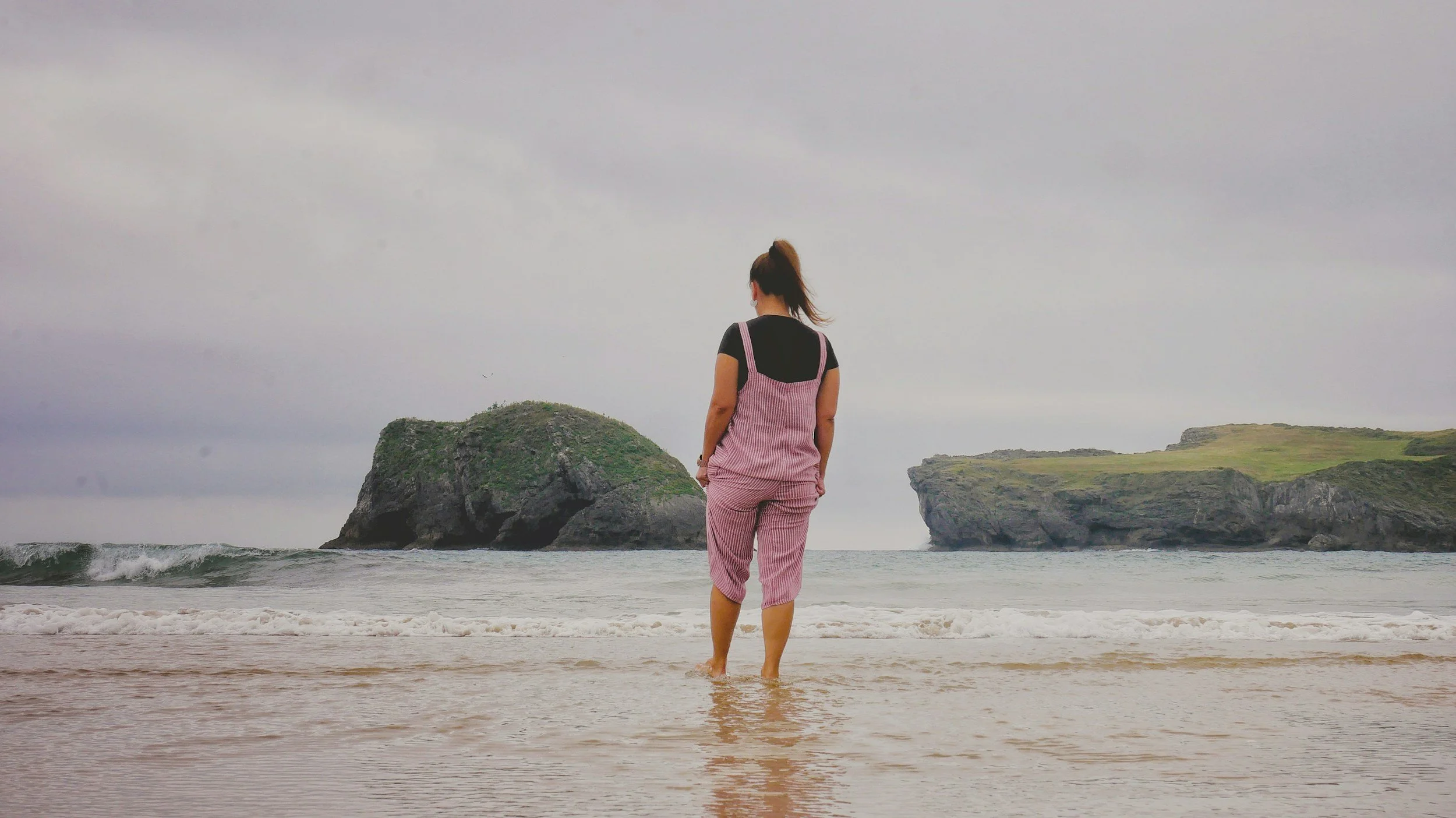 A woman standing ankle-deep in the ocean water facing towards large rocky islands under an overcast sky.