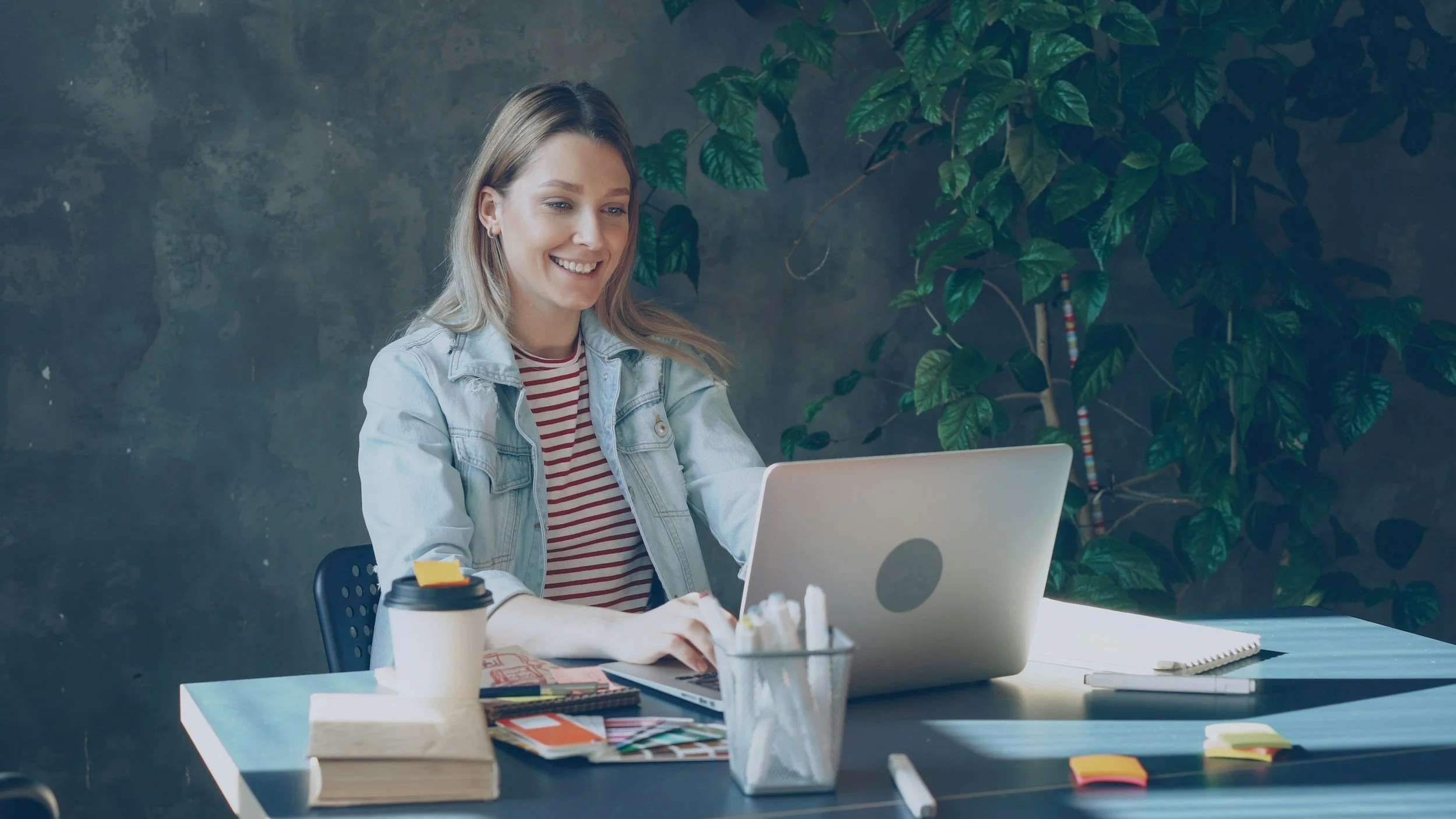 A woman with blonde hair wearing a light denim jacket and striped shirt working on a laptop at a desk decorated with notebooks, a coffee cup, sticky notes, pens, and a potted plant, in a modern office.