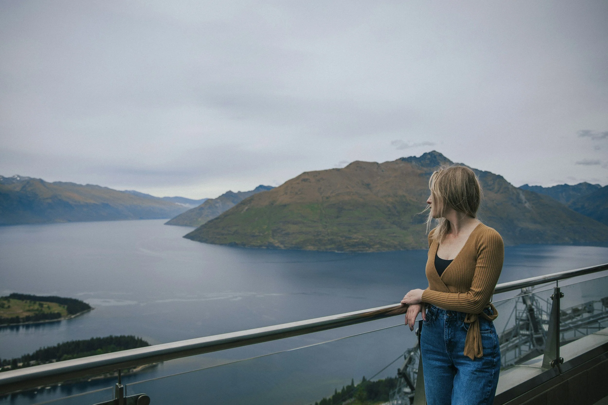 A woman with blonde hair wearing a brown ribbed sweater and blue jeans is standing at a railing overlooking a large body of water with mountains in the background on a cloudy day.