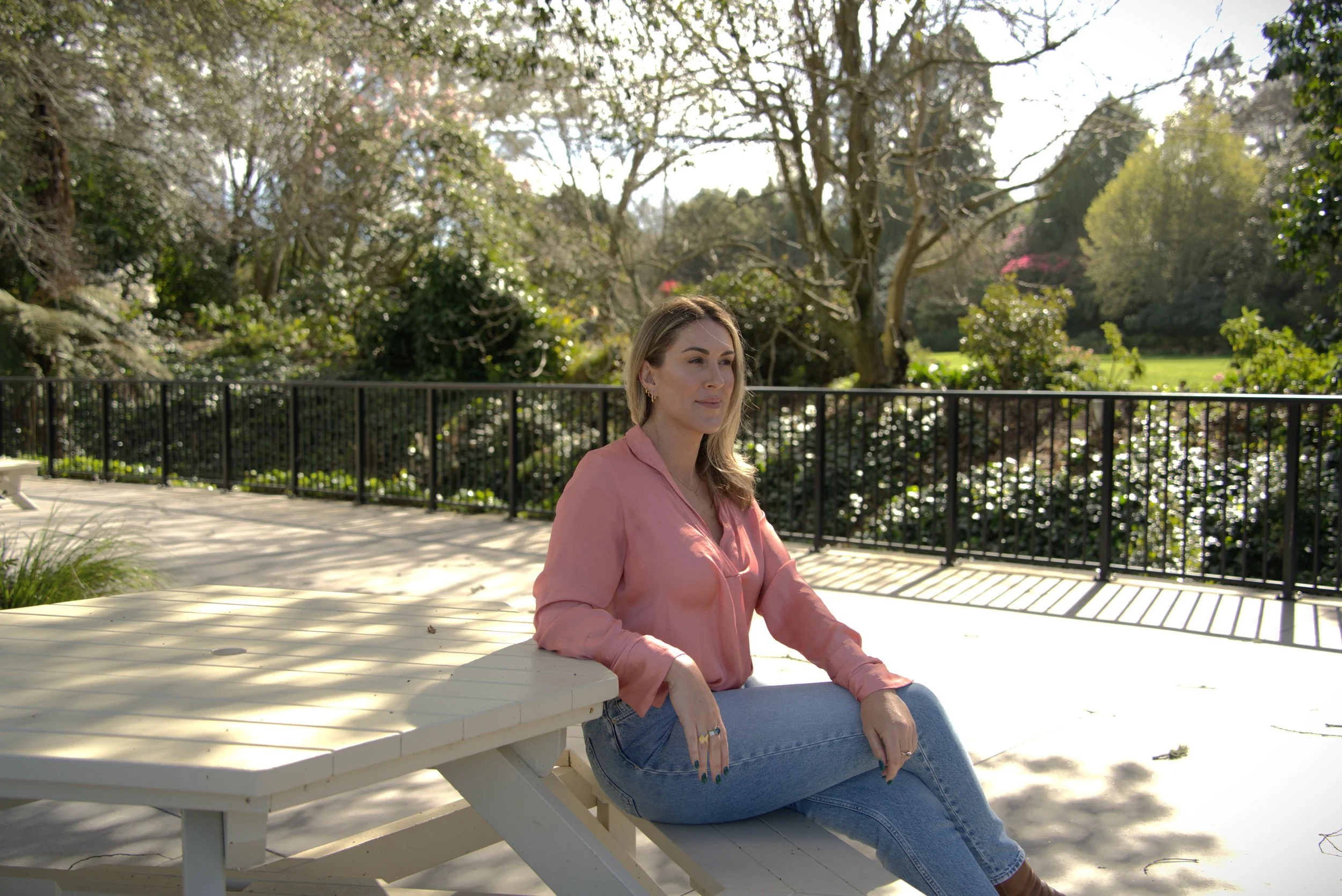 Genevieve Gray, RTT hypnotherapist and wellbeing coach based in Hamilton, New Zealand, in a pink blouse and jeans sitting outdoors at a white picnic table on a sunny day, with trees and greenery in the background.
