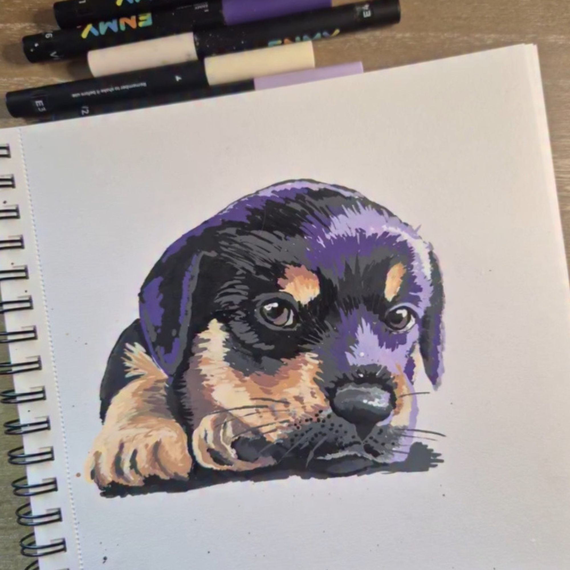 A spiral-bound notebook with a drawing of a rottweiler puppy on a white page, placed on a wooden surface, with several black and purple markers nearby.