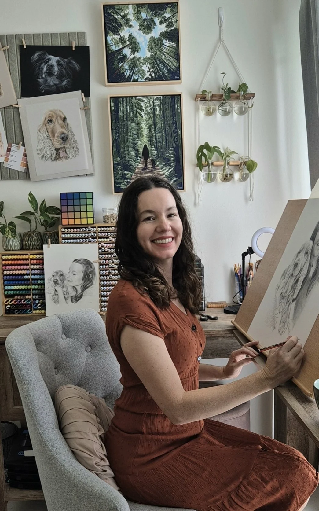 A woman with dark curly hair, wearing a rust-colored dress, is sitting in a light grey upholstered chair in an art studio. She is smiling while holding a pencil and working on a detailed animal portrait, on a canvas. The studio has shelves filled with colored pencils and sketches of dogs hanging on the wall, along with two landscape paintings and potted plants, creating a cozy and creative atmosphere.
