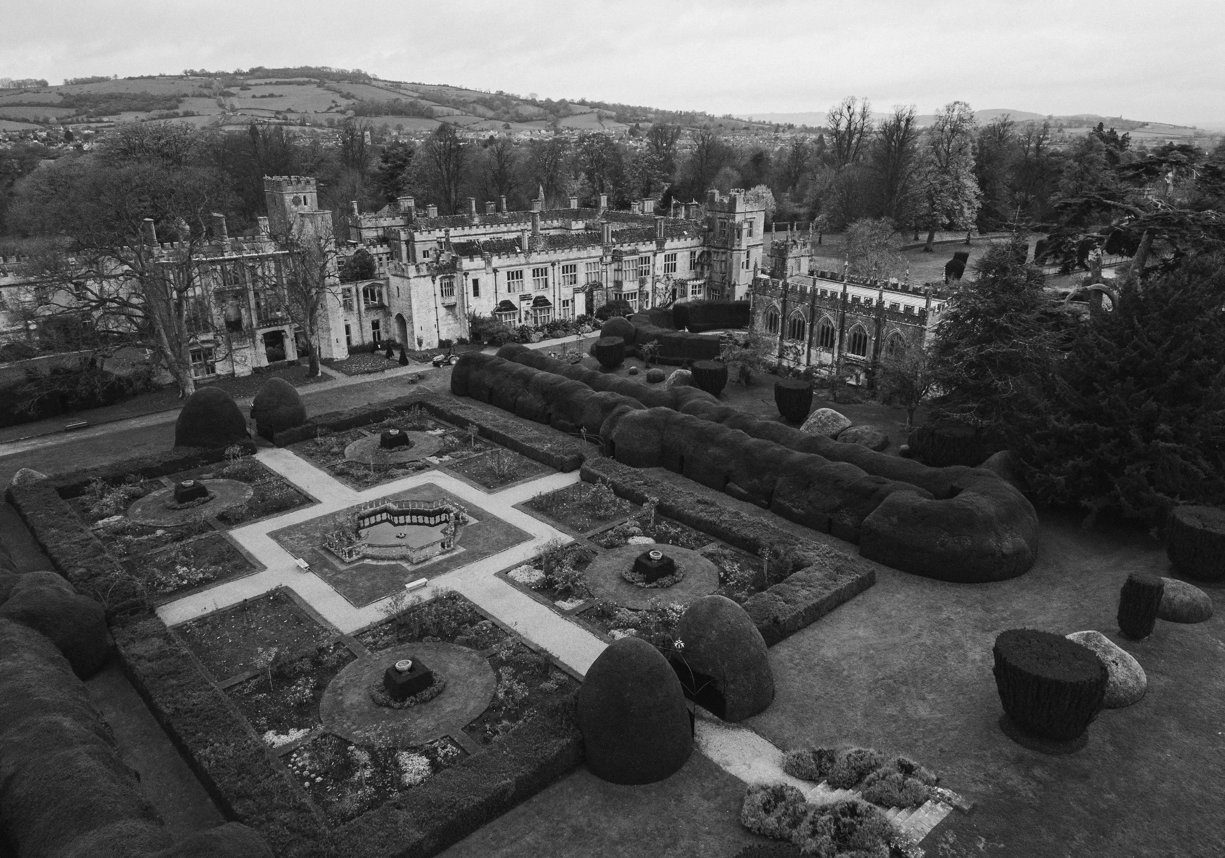 Black and white aerial view of a castle with landscaped gardens and pathways, surrounded by trees and rolling hills in the background at sudeley castle, Gareth roy photography