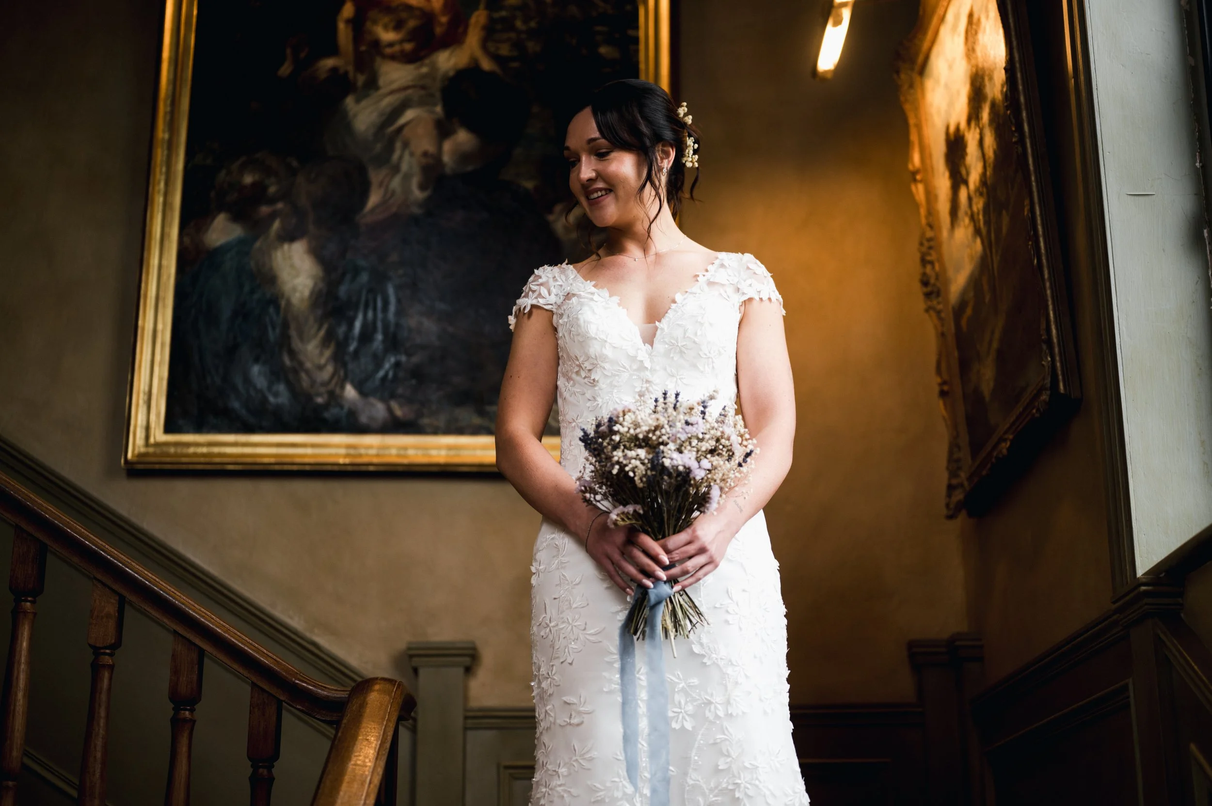 A bride in a white wedding dress holding a bouquet of flowers, standing on a staircase in front of a dark background with framed artwork pauntley court, Gareth roy photography
