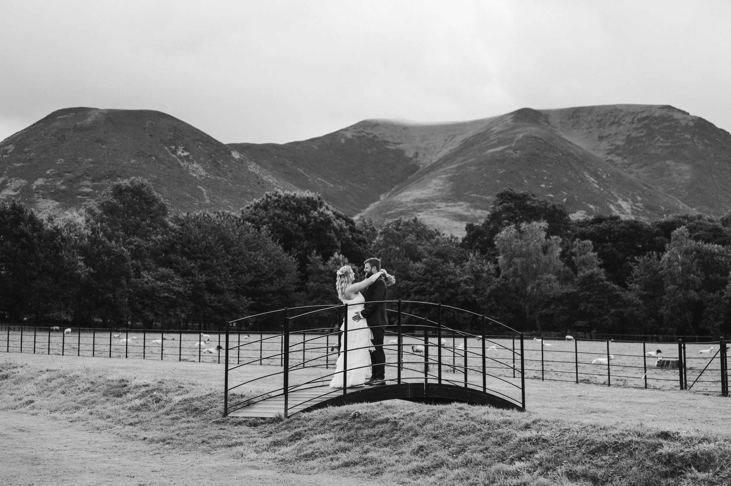 A black-and-white photo of a bride and groom standing on a small bridge, embracing and looking at each other, with mountains and trees in the background.