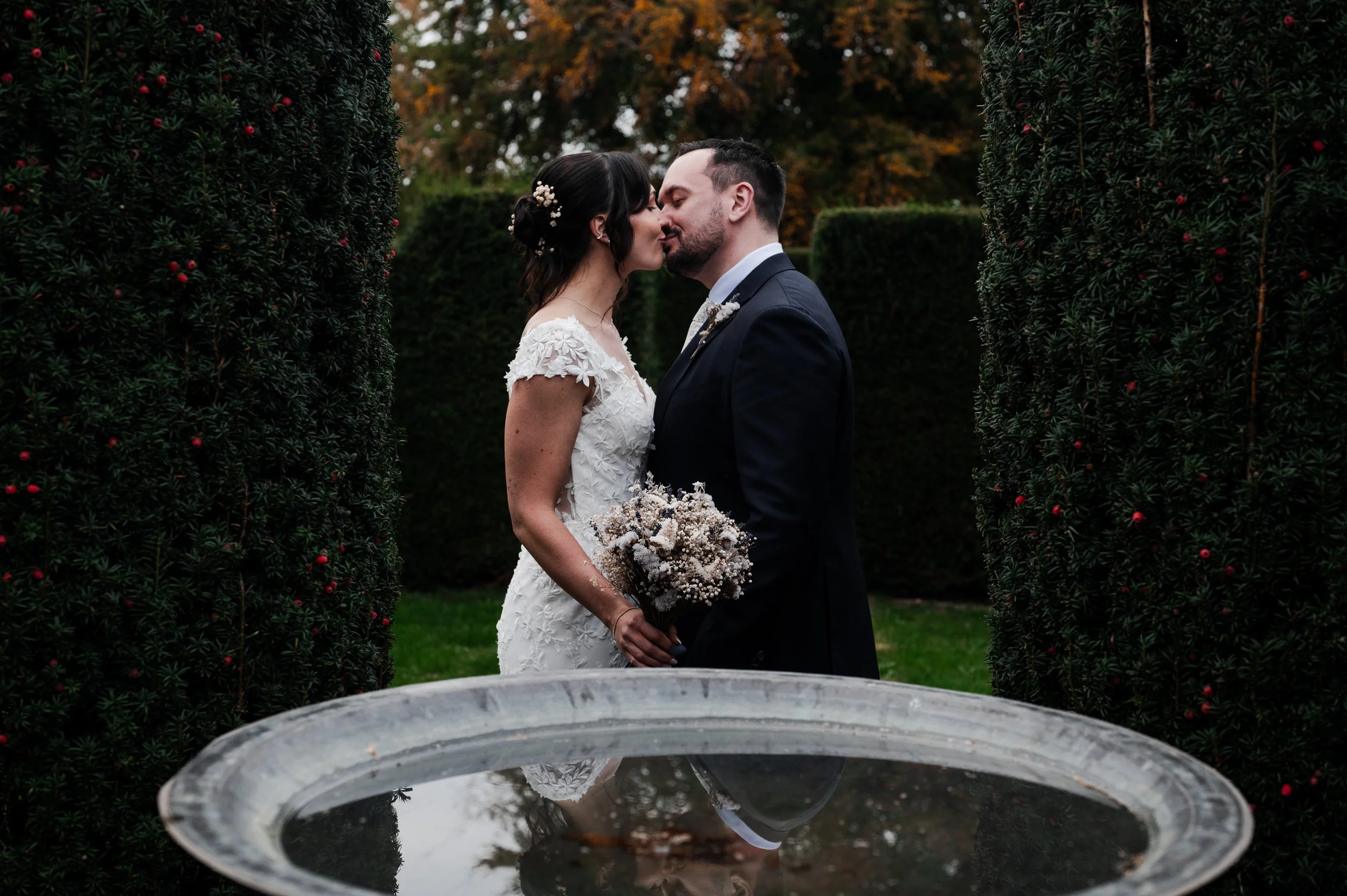 A bride and groom sharing a kiss on their wedding day, standing between tall evergreen bushes with red berries, reflected in a fountain in the foreground at sudeley castle, Gareth roy photography