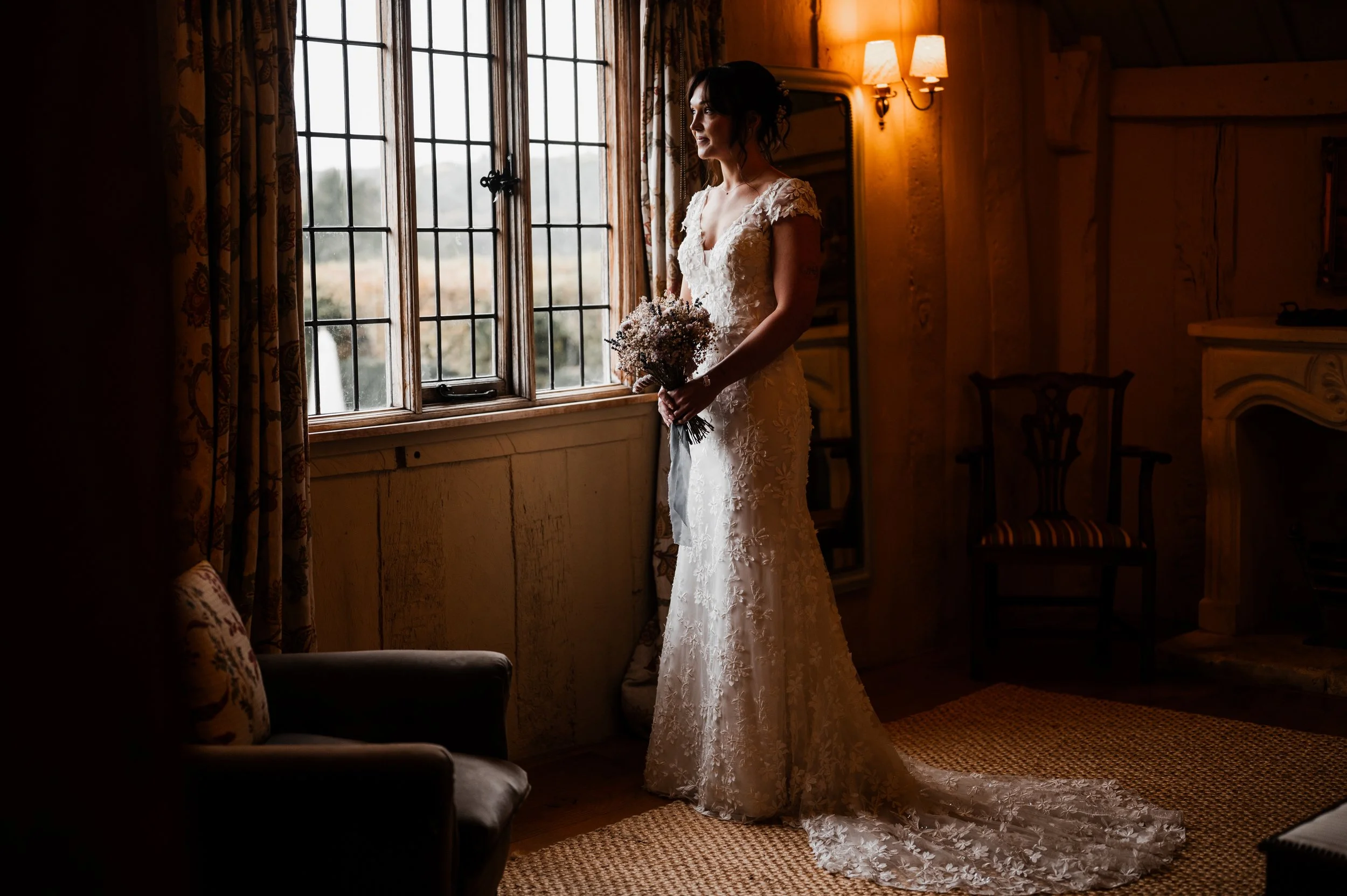 A bride in a lace wedding dress holding a bouquet, standing by a window with wooden frame and patterned curtains in a warmly lit room pauntley court, Gareth roy photography