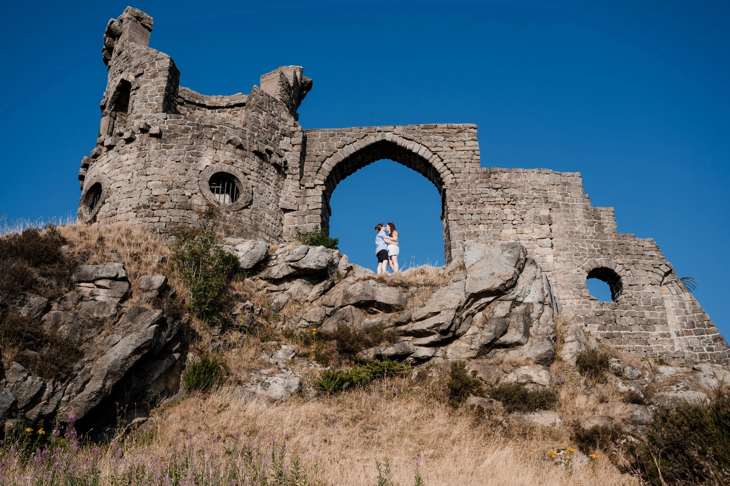 Mow Cop Engagement Shoot - two women stood under an arch of a castle ruin while photographer looks up the hill. 