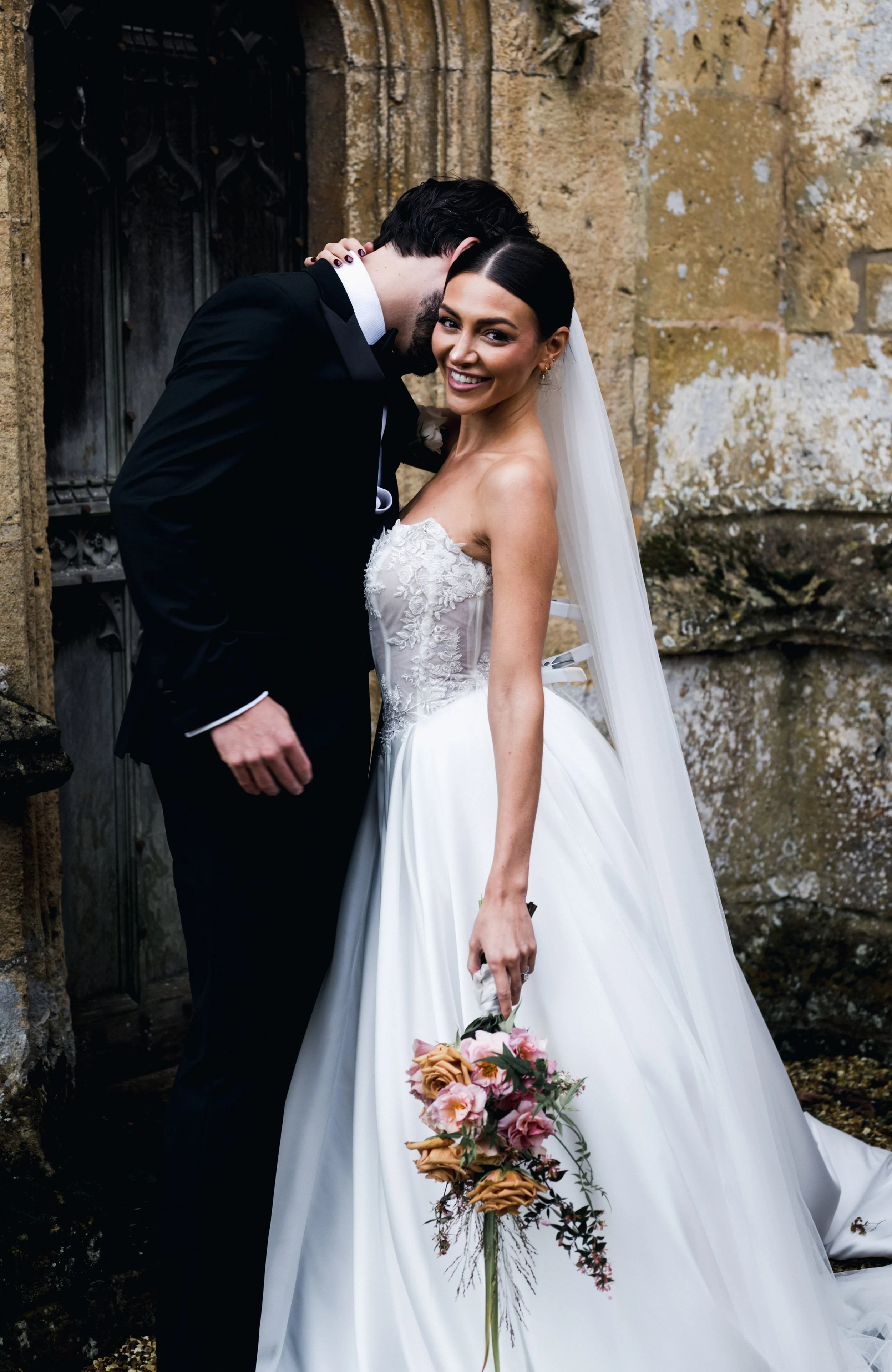 A bride in a white wedding dress smiling and holding a bouquet of pink and peach flowers, standing outdoors near a stone wall while a groom in a black tuxedo whispers to her.