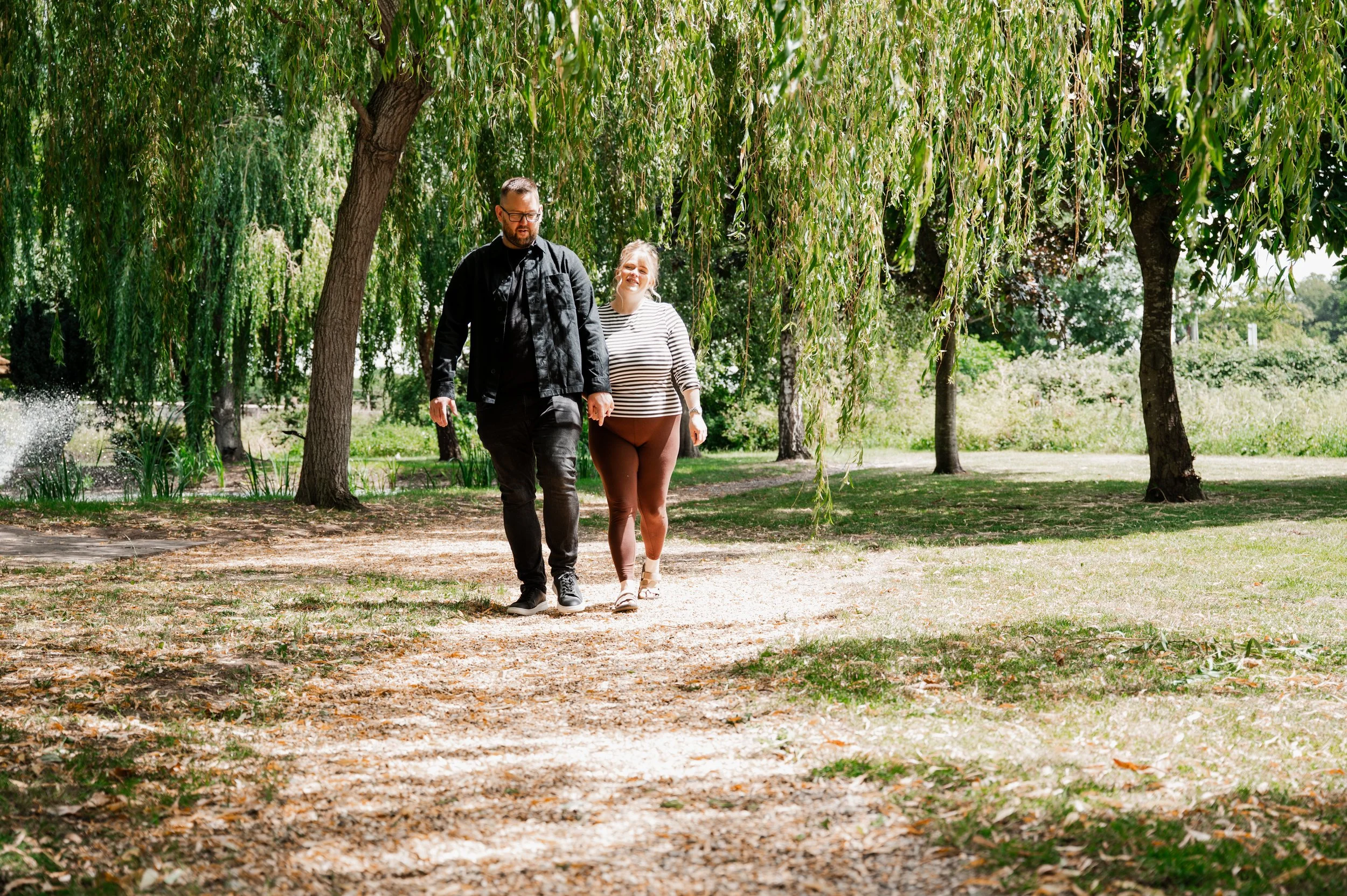 The Grosvenor Pulford Engagement - man in black shirt and woman in striped top, walking down a path and smiling, surrounded by trees