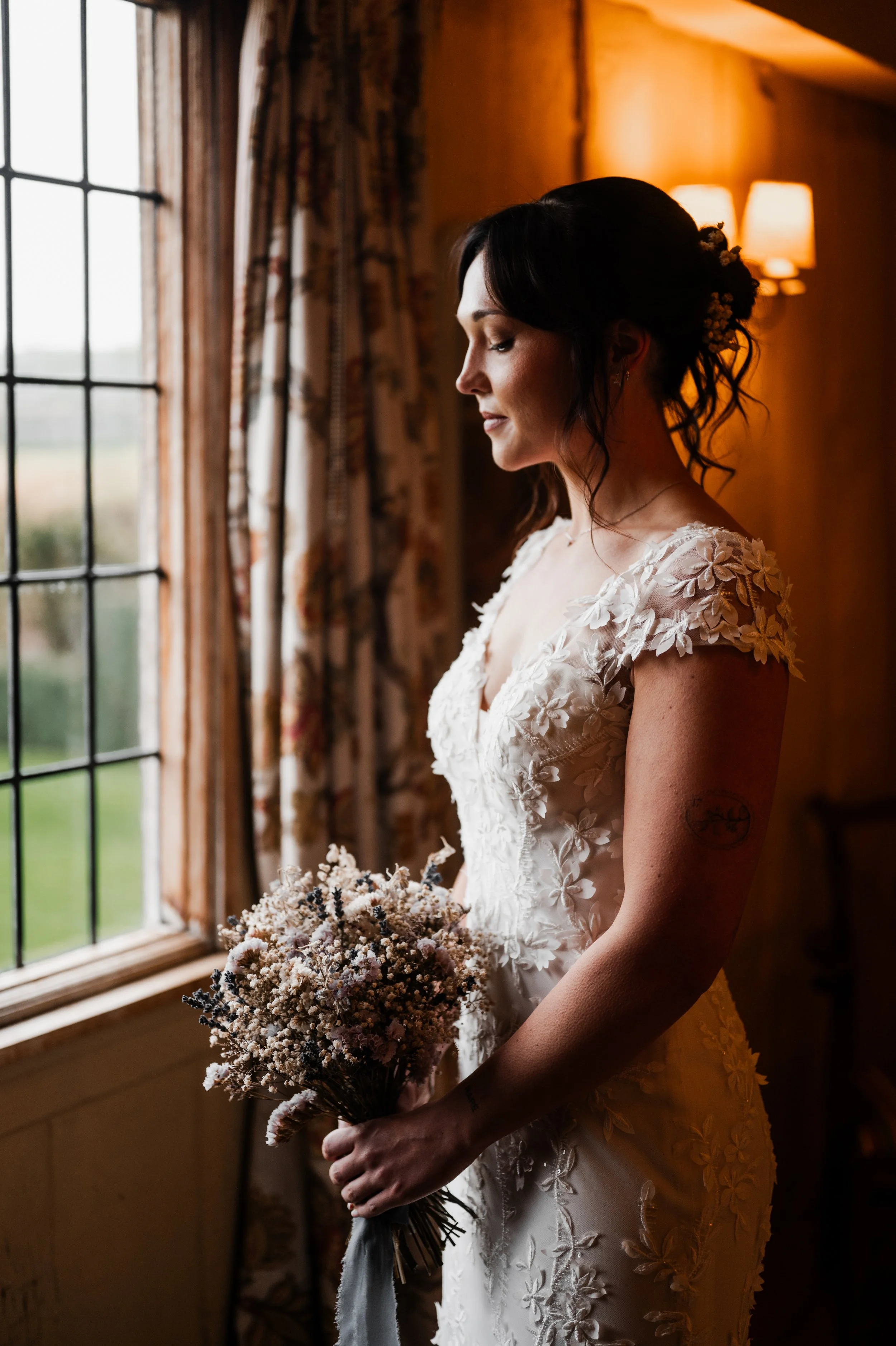Bridal woman in a wedding dress holding a bouquet, standing indoors near a window with curtains and warm lighting pauntley court, Gareth roy photography