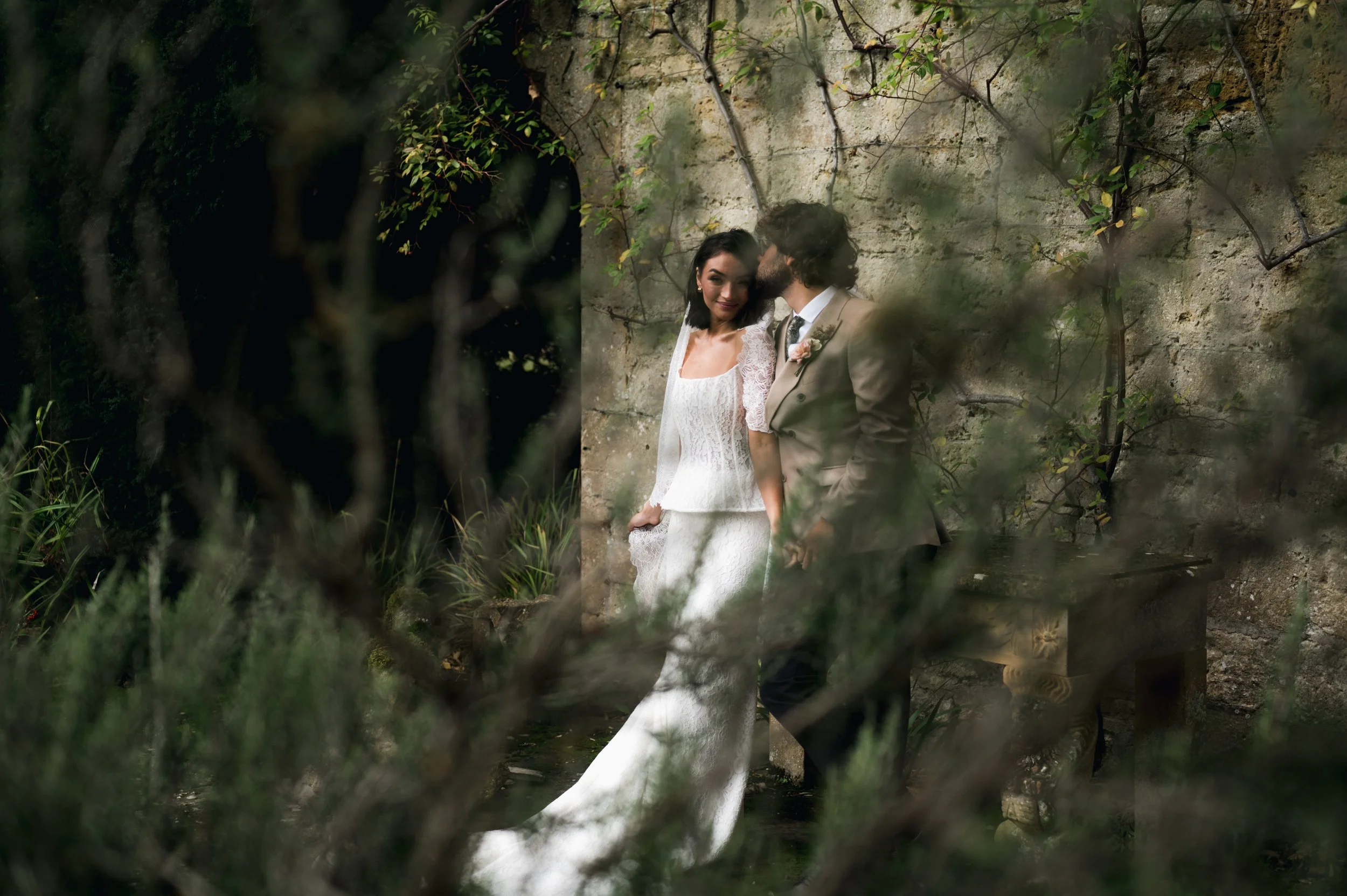 A bride and groom standing close together against a rustic stone wall, with greenery and branches partially obscuring the view at sudeley castle, Gareth roy photography